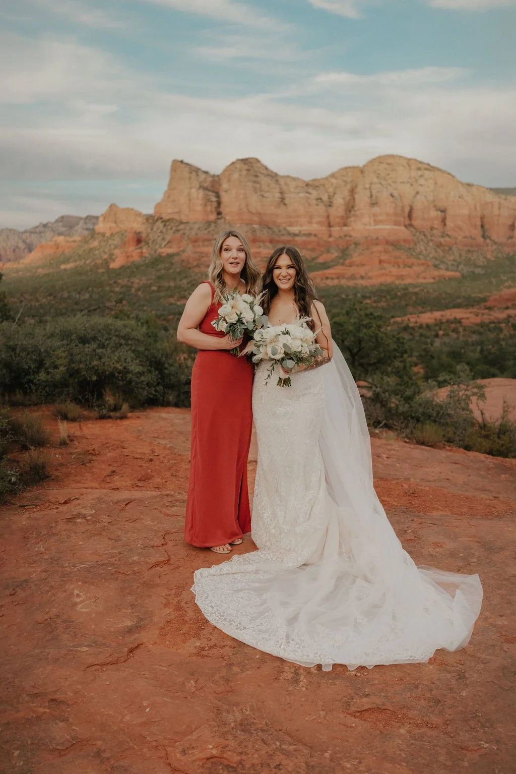Two women, one in a white wedding dress and the other in a red gown, standing outdoors on a rocky terrain with red sandstone formations in the background, holding bouquets of flowers, smiling at the camera during sunset.