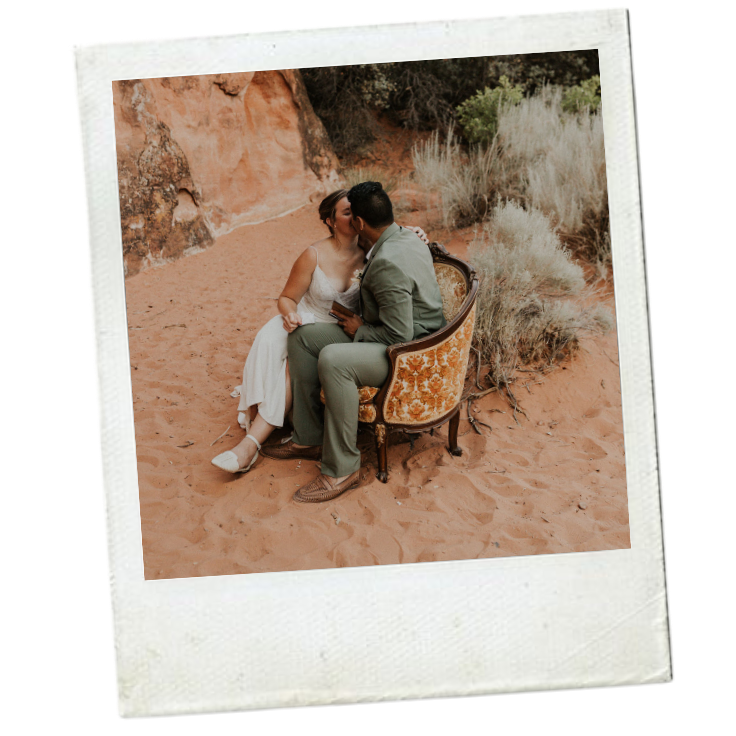 A couple sits on a vintage armchair in a desert landscape, sharing a kiss.