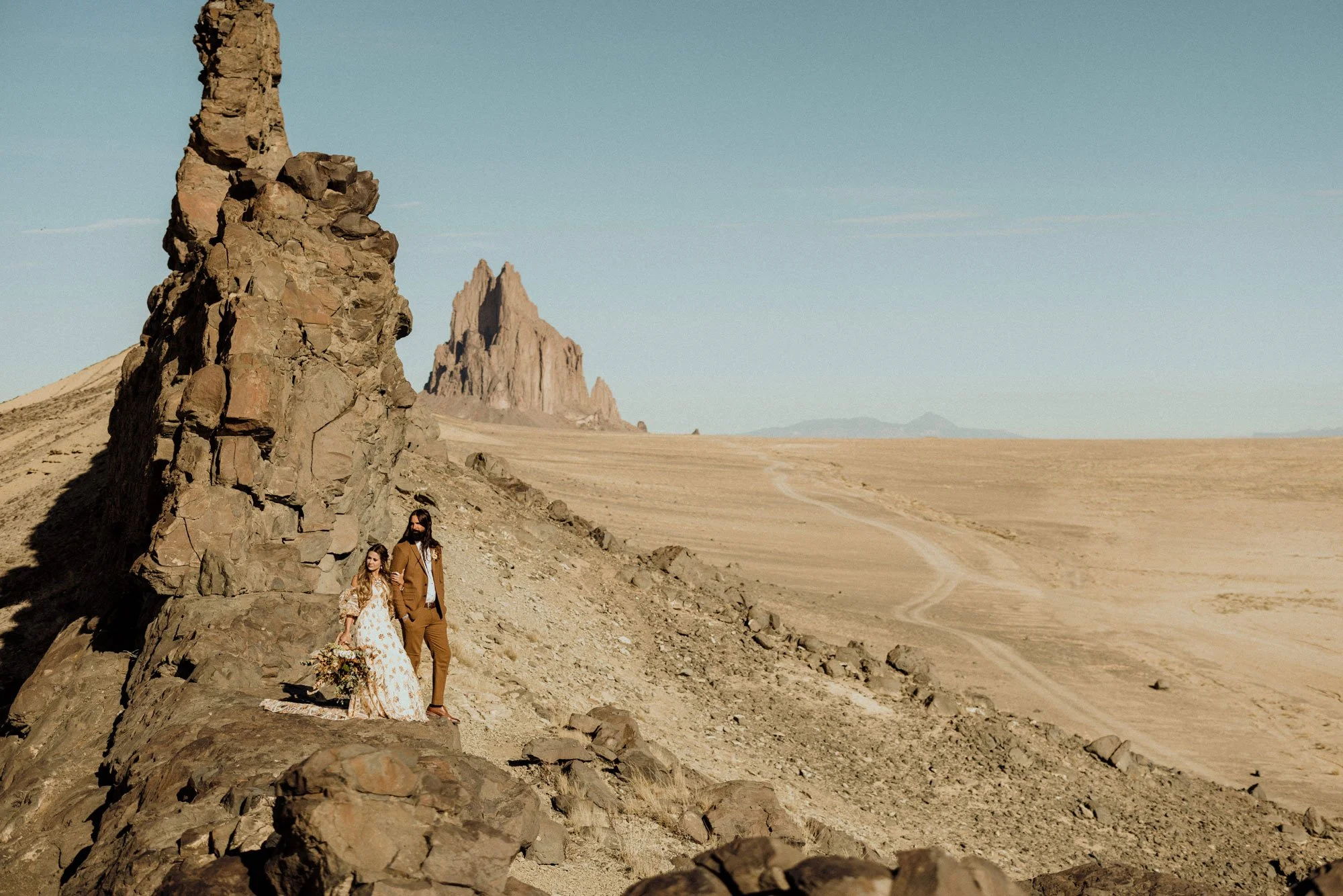 A couple in wedding attire standing on rocky terrain in a desert landscape with large rock formations and mountains in the background.