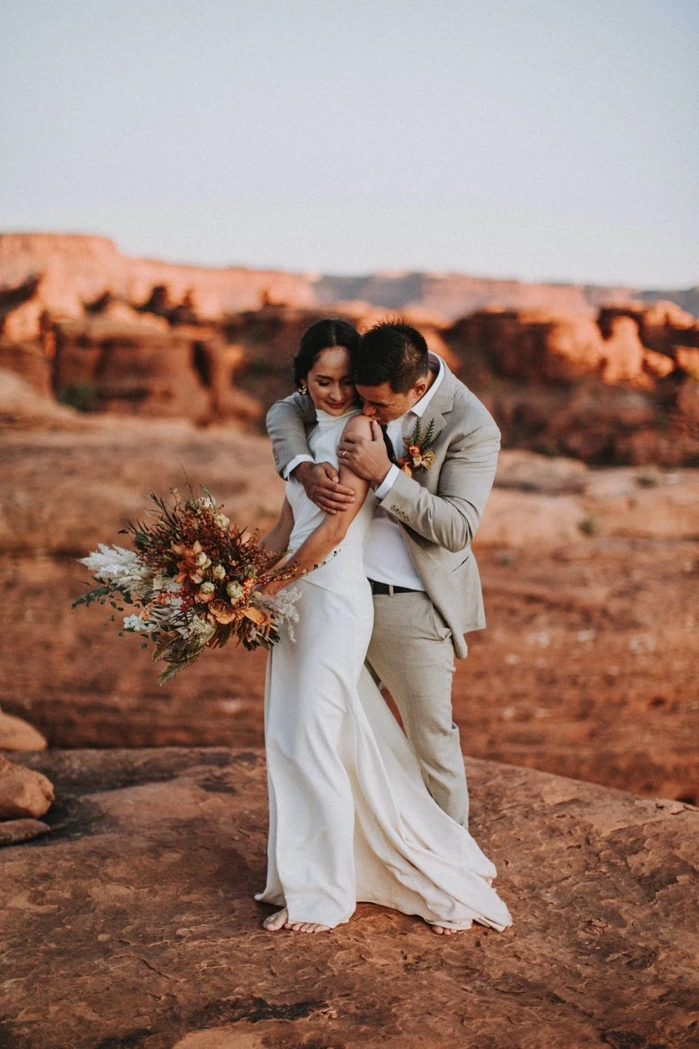 A bride and groom share a joyful moment outdoors in a desert landscape, with the bride holding a bouquet of flowers and both embracing.