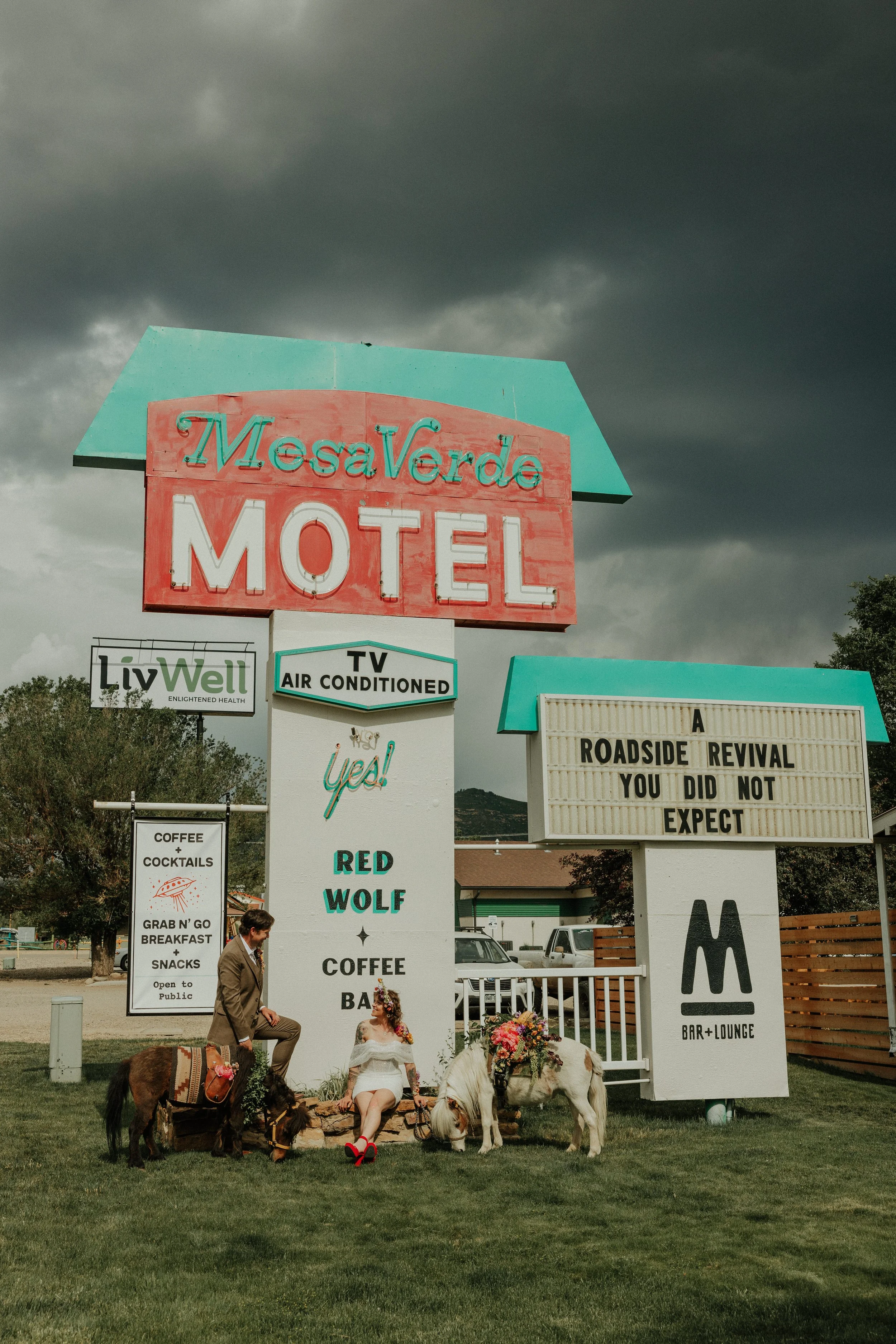 A vintage motel sign for Mesa Verde Motel with a dark clouded sky in the background. Two small ponies with flower decorations are sitting on the grass, accompanied by a man in a suit and a woman in a white dress with red shoes.