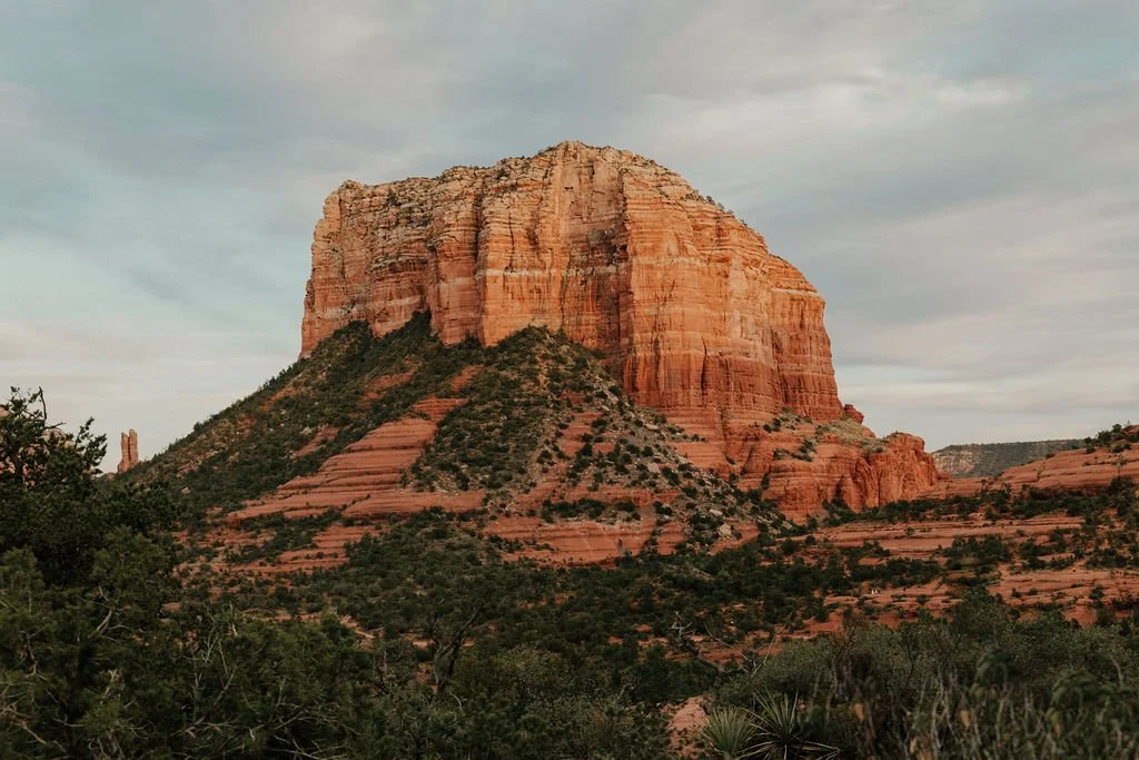 A large red sandstone mesa with layered rock formations, surrounded by green shrubbery and a cloudy sky in the background.