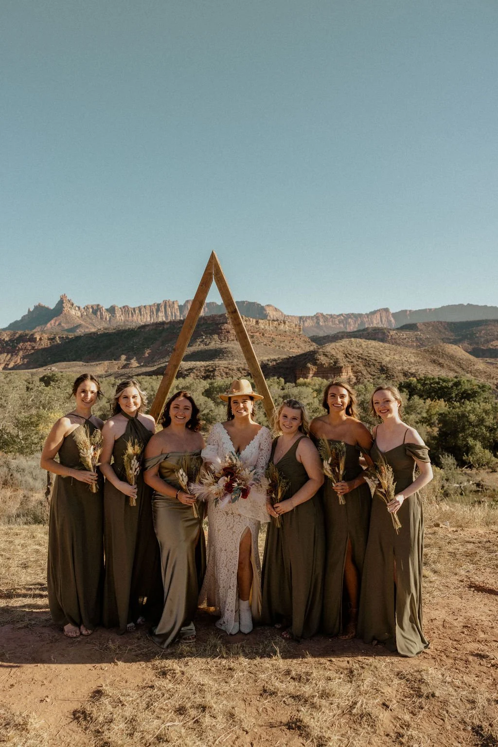 A bride and six bridesmaids standing outdoors in a desert landscape with mountains in the background, holding bouquets, with a wooden triangular arch behind them.