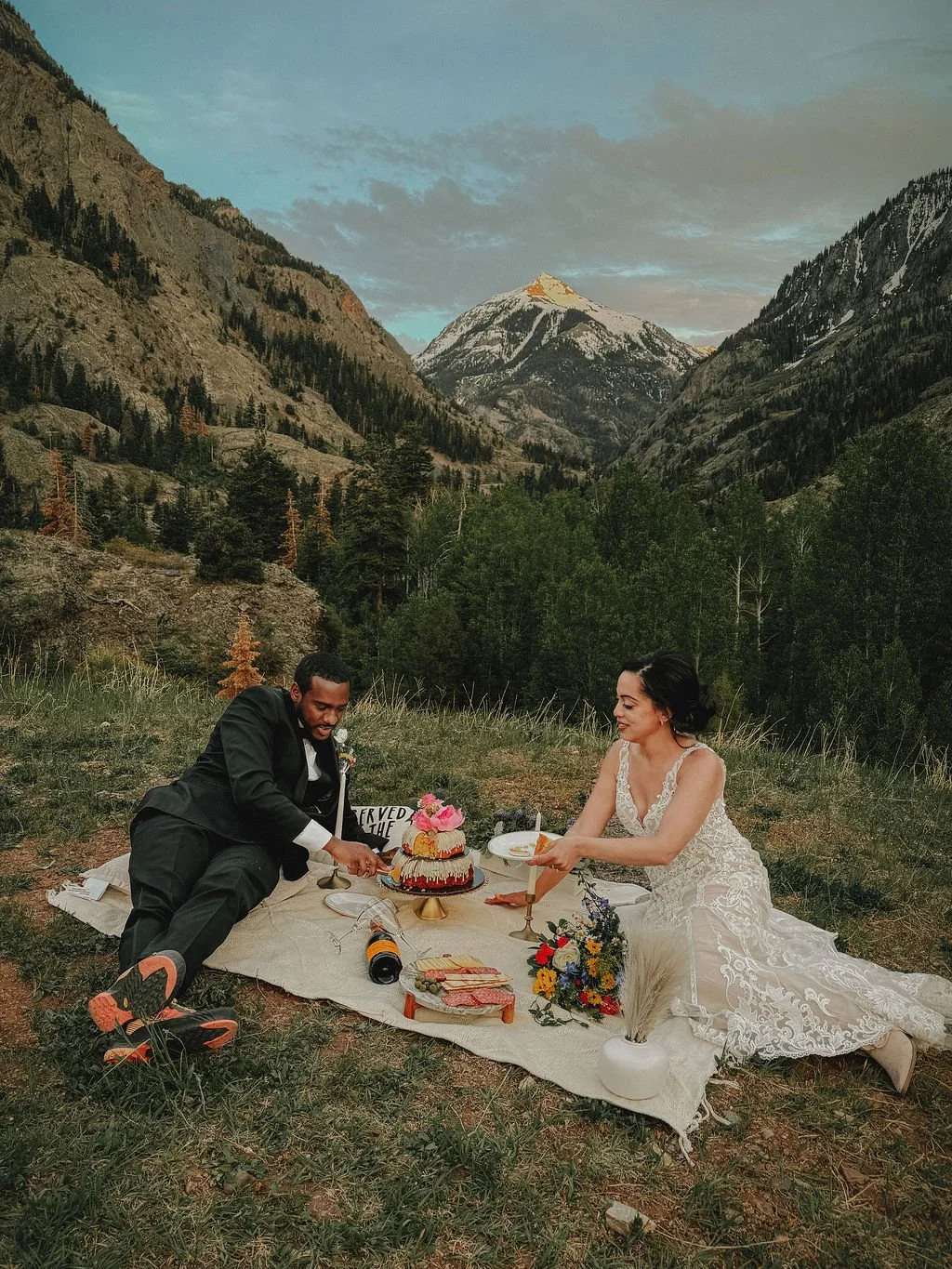 A man and woman in wedding attire having a picnic with a wedding cake outdoors in a mountainous landscape with tall trees, grassy fields, and snow-capped peaks in the background.