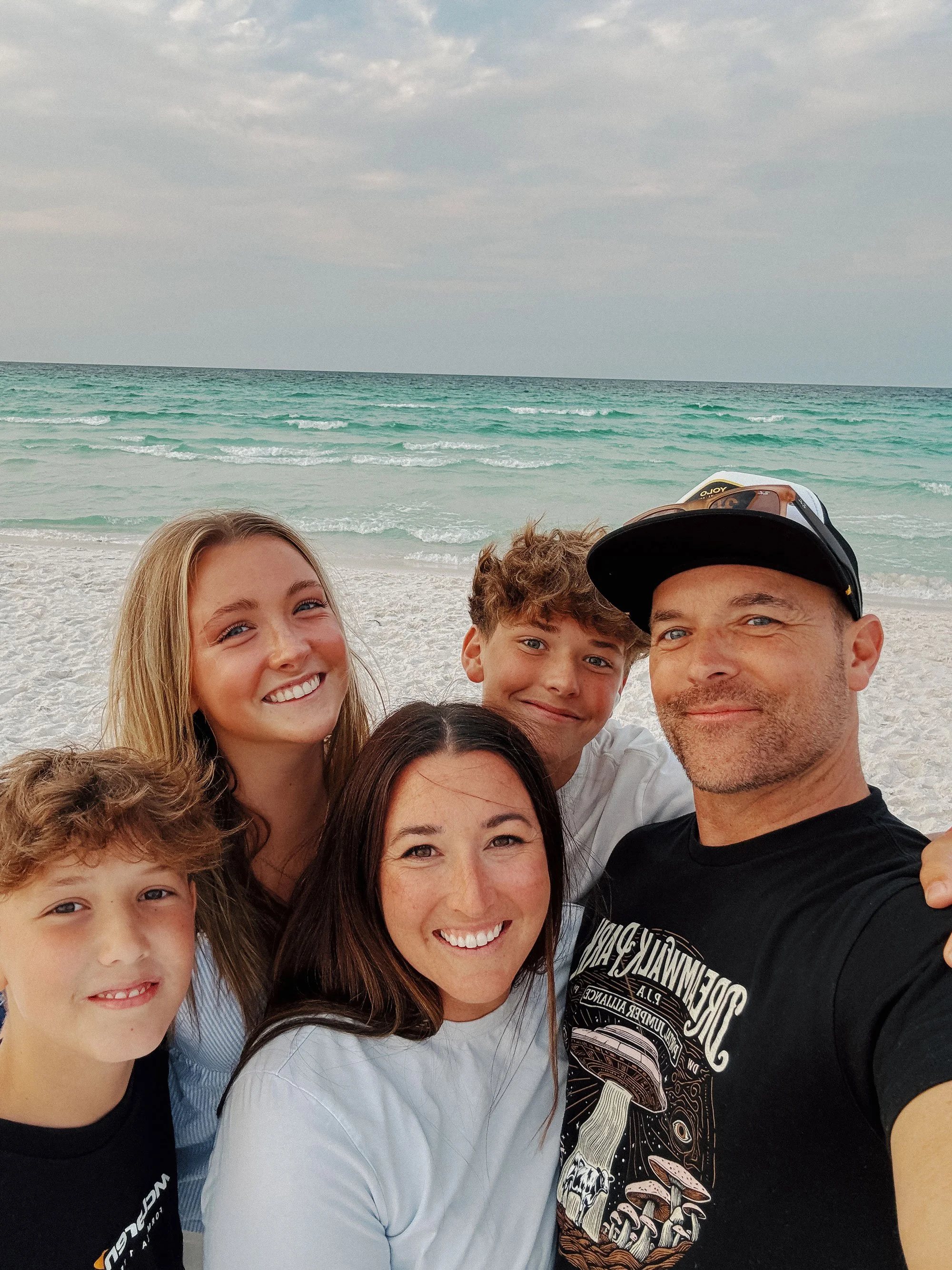 Family of five taking a selfie on the beach near the ocean, with waves and cloudy sky in the background.