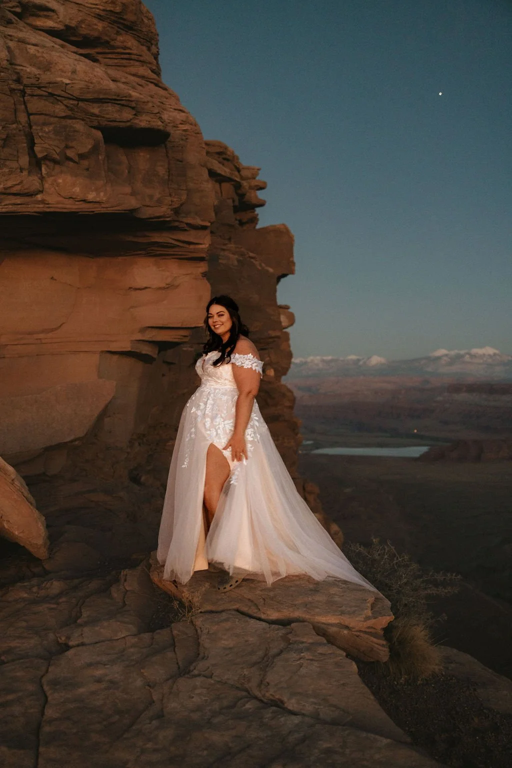 A woman in a wedding dress standing on a rocky ledge beside a large red rock formation at dusk with a mountain landscape and a river in the background.