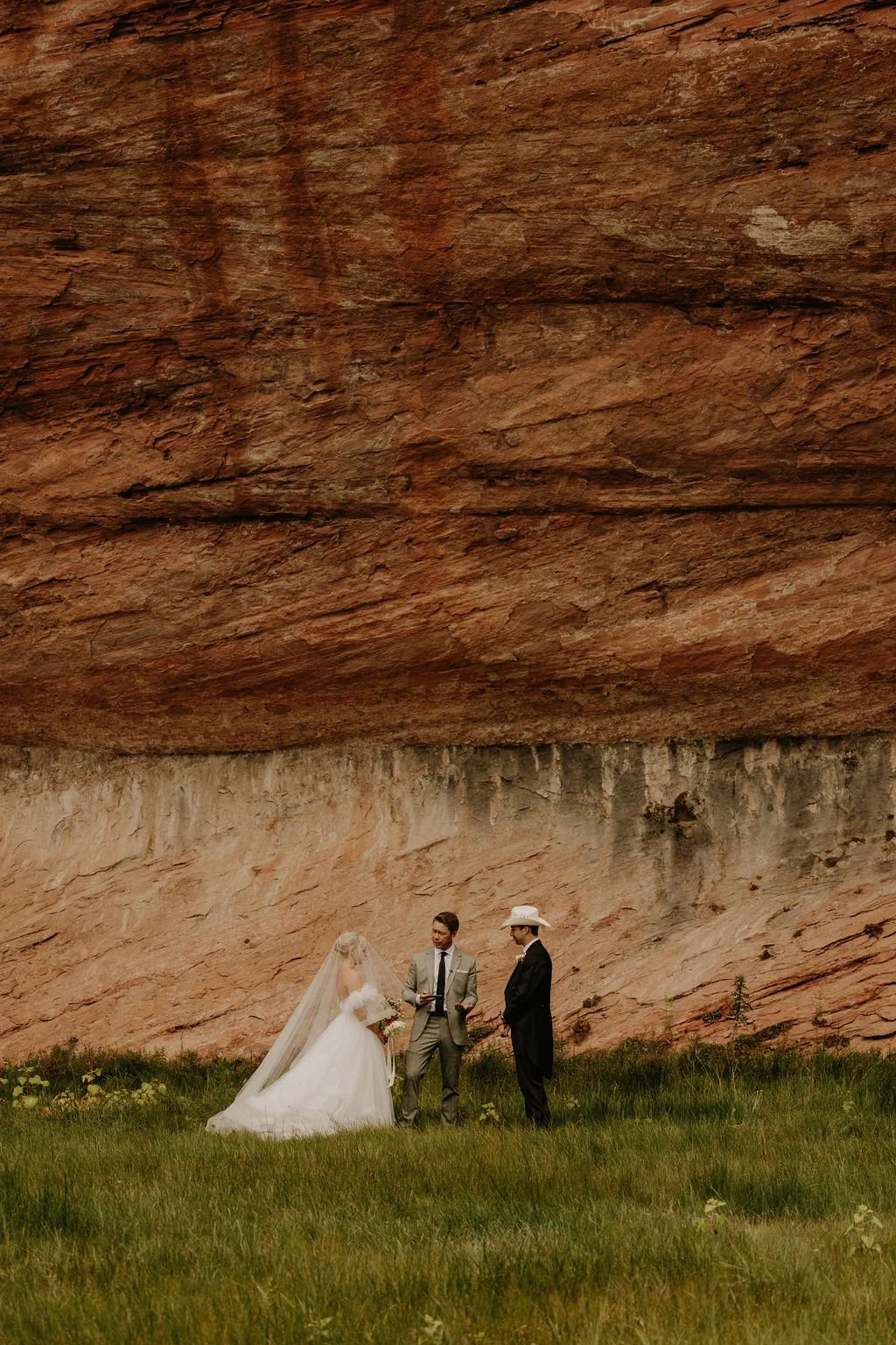 A wedding ceremony with a bride, groom, and officiant standing on green grass in front of a large reddish rock formation.