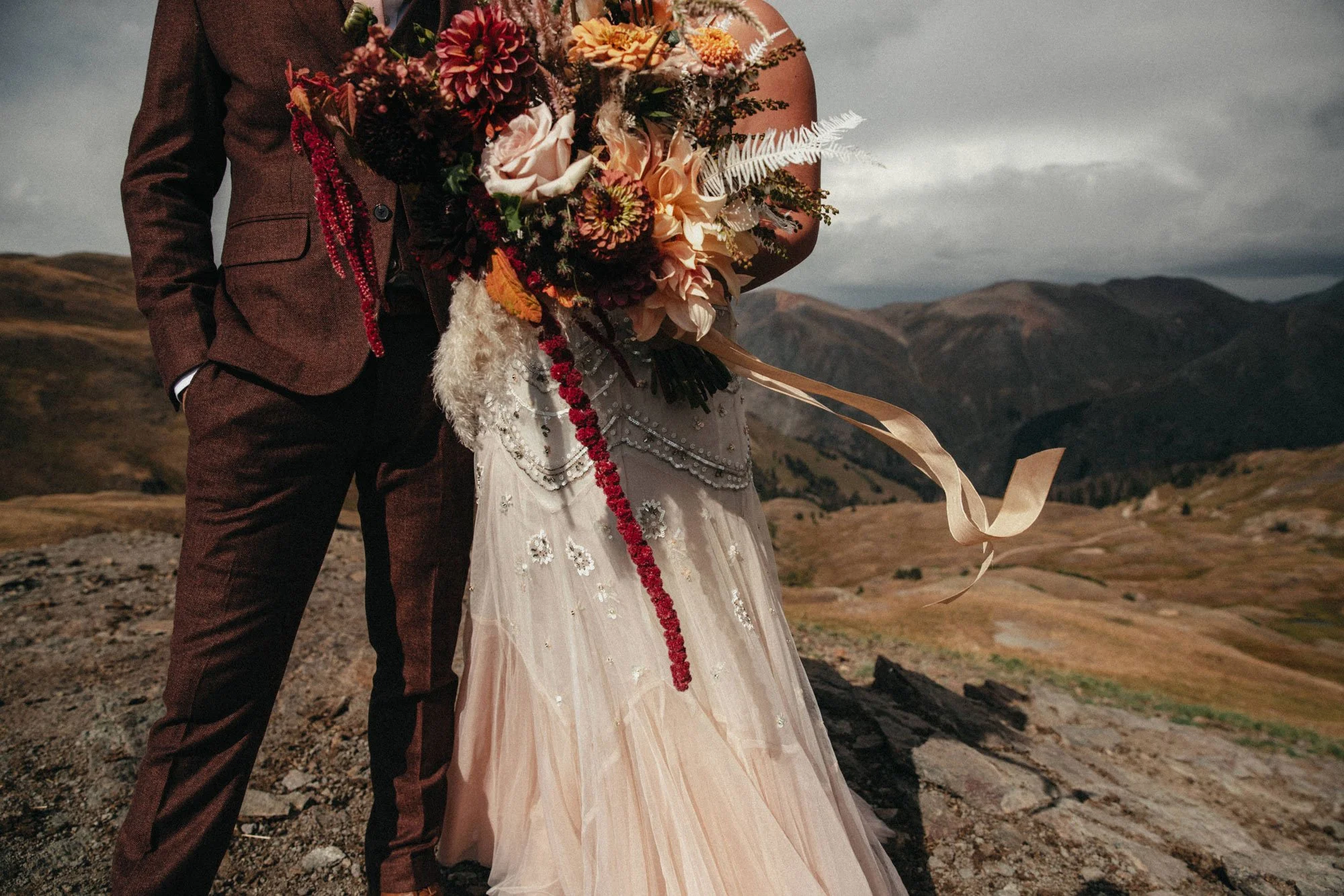 A couple dressed in formal attire standing outdoors on rocky terrain with mountains in the background, holding a bouquet of flowers.