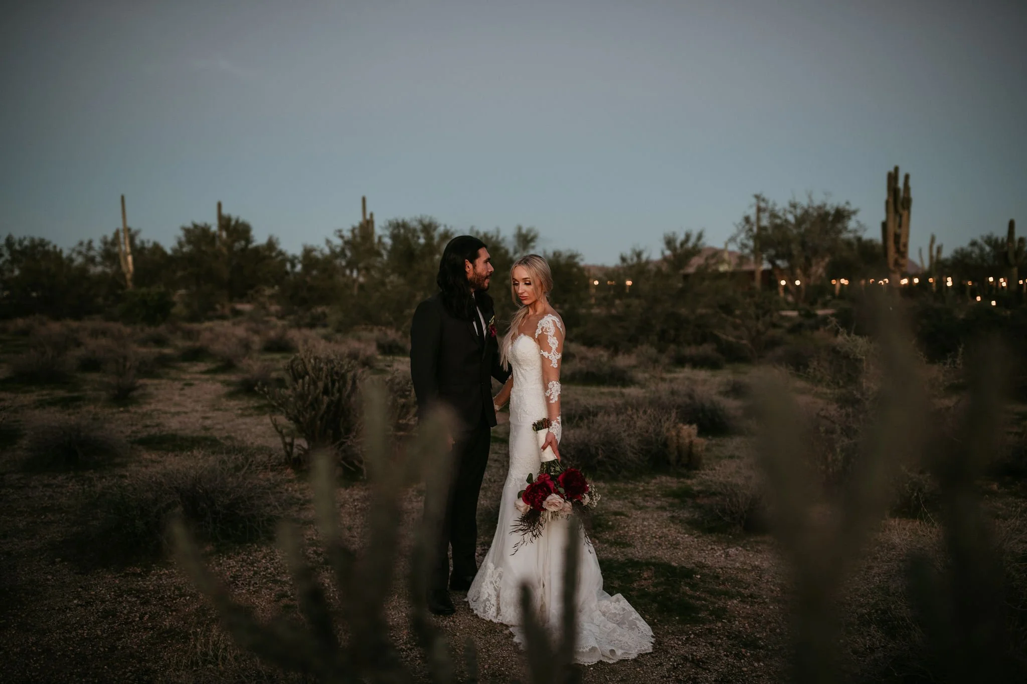 A bride and groom stand close together at sunset in a desert landscape with cacti and sparse vegetation.