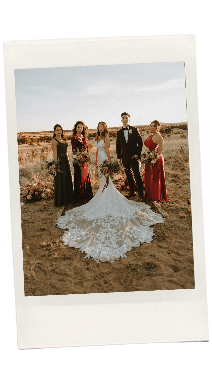 A wedding party standing outdoors on desert terrain during sunset, with the bride in a white lace wedding gown and the groom in a black tuxedo, surrounded by three women in colorful dresses.