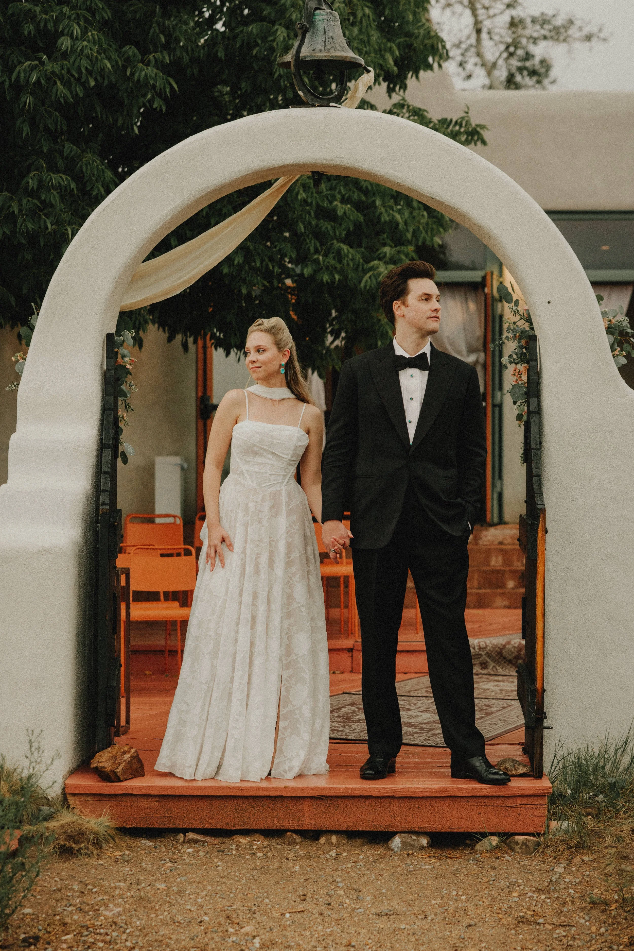 A bride and groom in wedding attire standing in a field, embracing each other, with a scenic landscape of rolling hills and trees in the background.