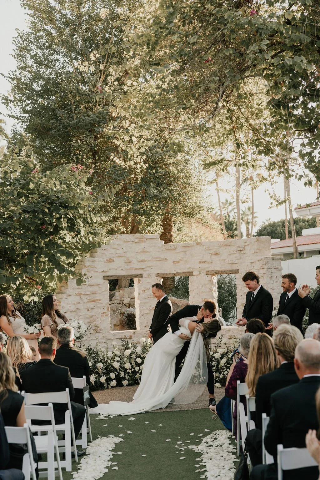 A wedding ceremony outdoors with the bride and groom sharing a kiss during their vows, surrounded by seated guests and natural greenery, with the bride dressed in a white gown and veil, and the groom in a black suit, in front of a white stone backdro