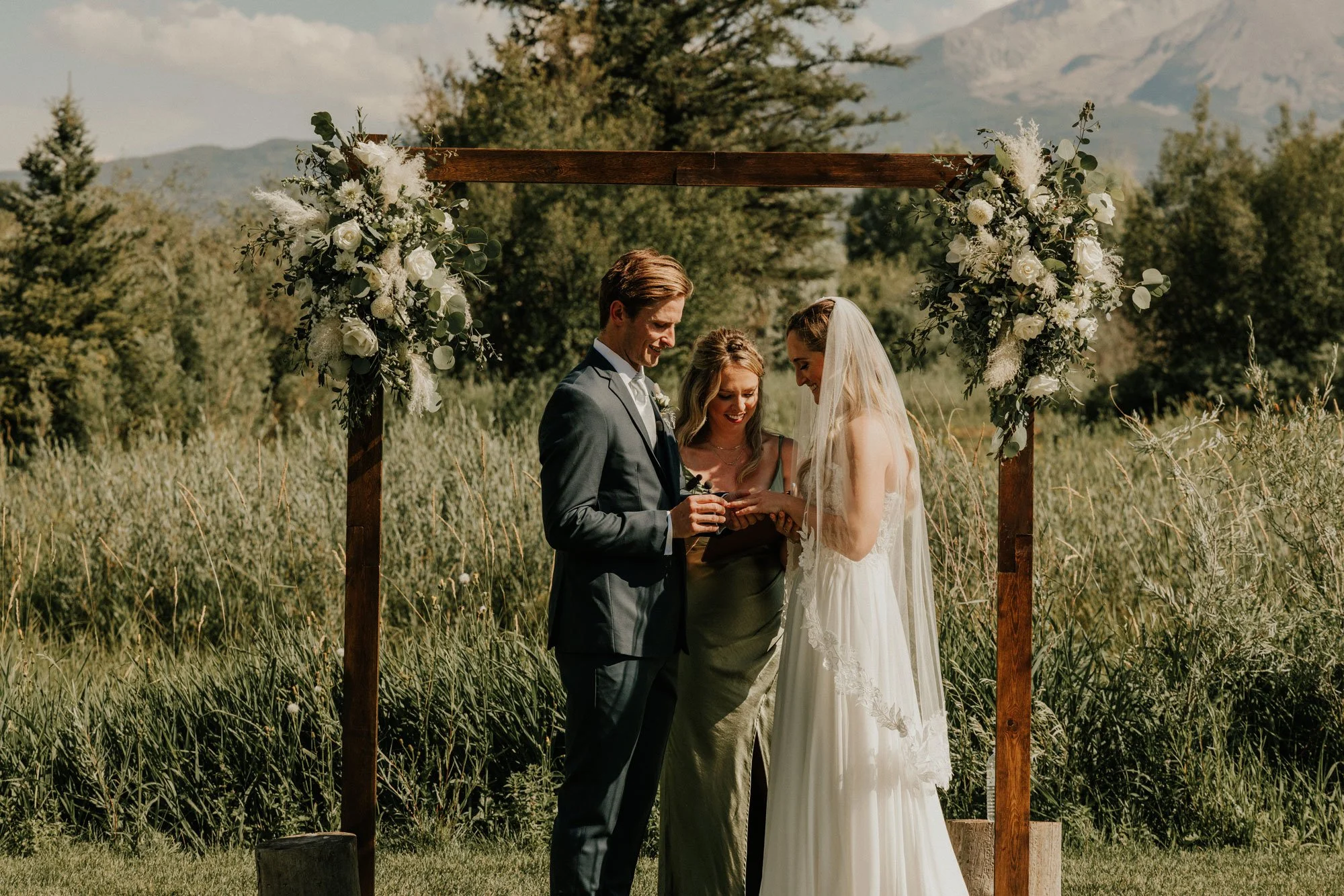 A couple getting married standing under a wooden arch decorated with white flowers and greenery, outdoors with mountains and trees in the background.