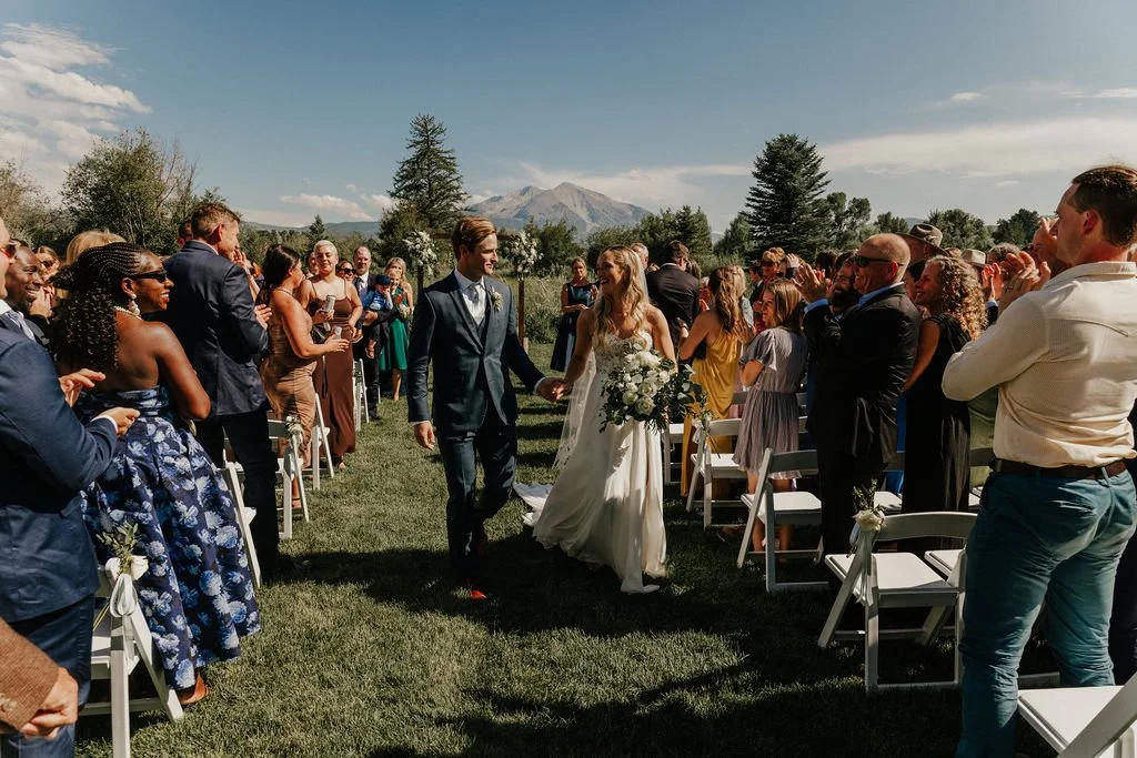 A bride and groom walking down the aisle at an outdoor wedding ceremony with guests clapping and smiling, mountains and trees in the background.