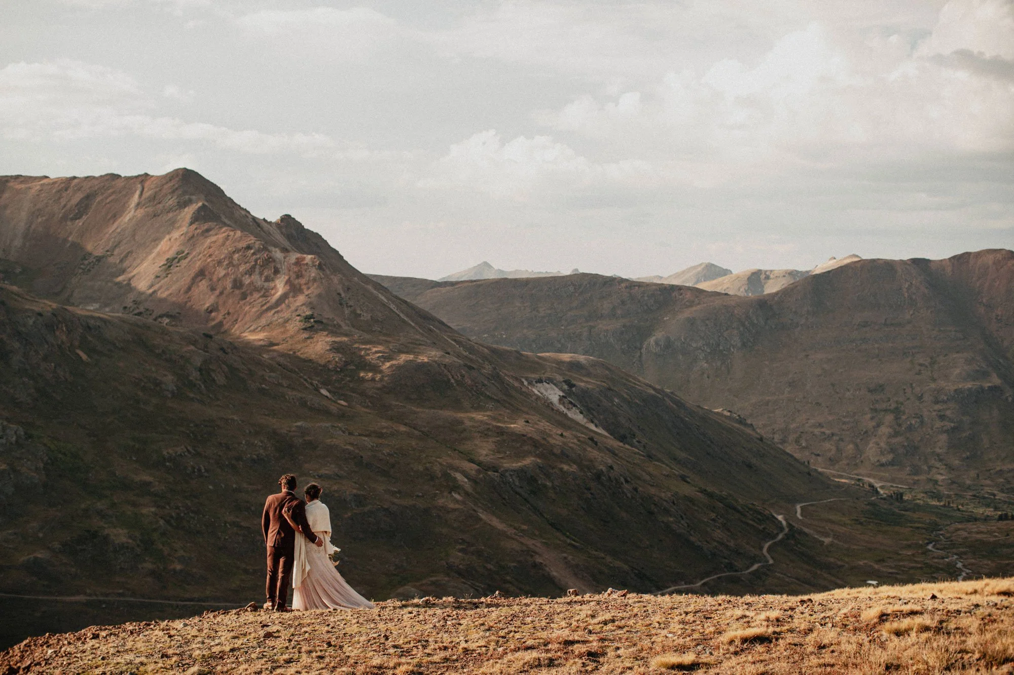 A couple, a man and a woman, standing on a grassy hilltop holding hands with mountains and a cloudy sky in the background.