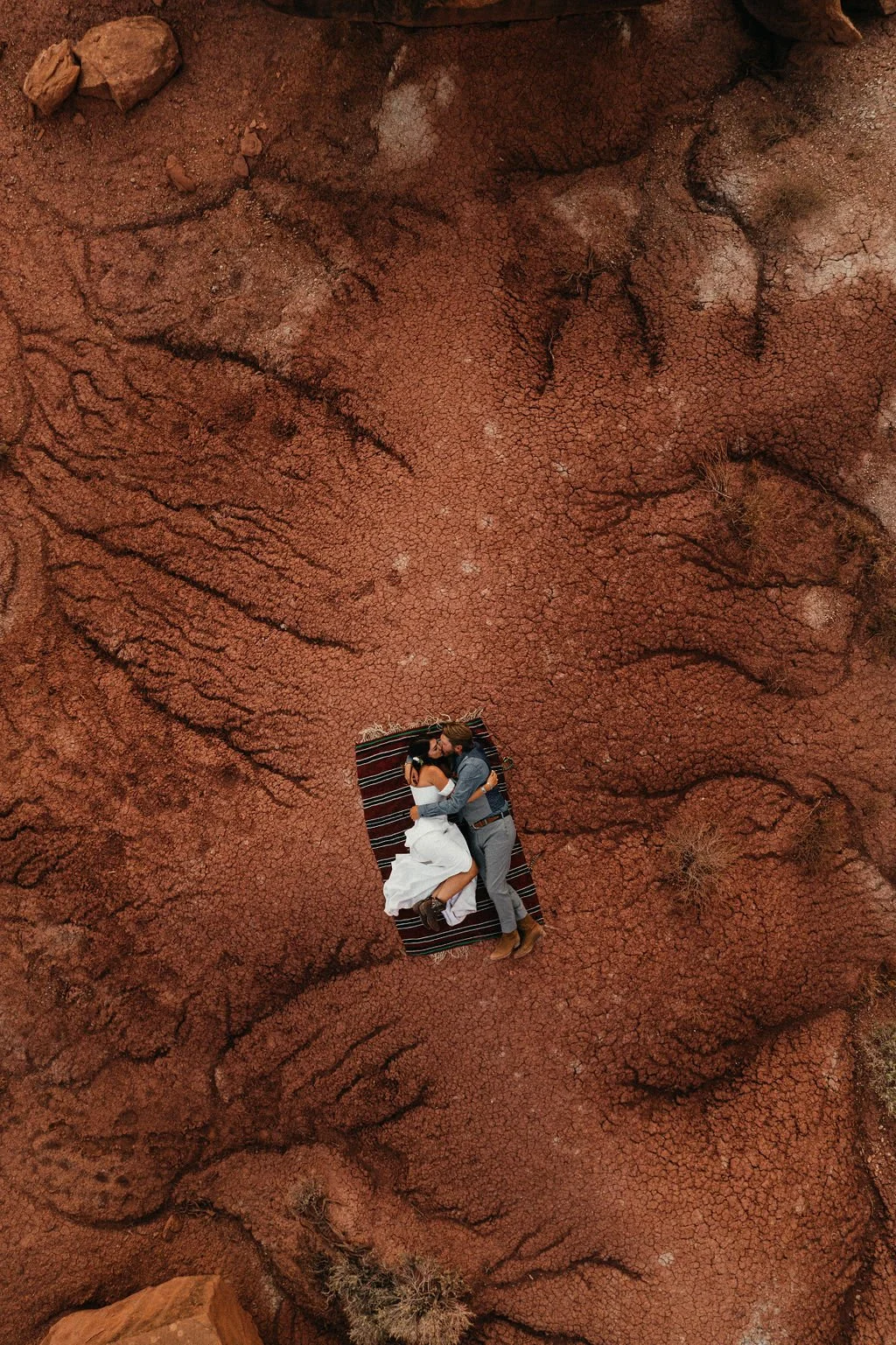 An aerial shot of a couple lying on a striped blanket on cracked, reddish-brown desert ground, embracing each other.