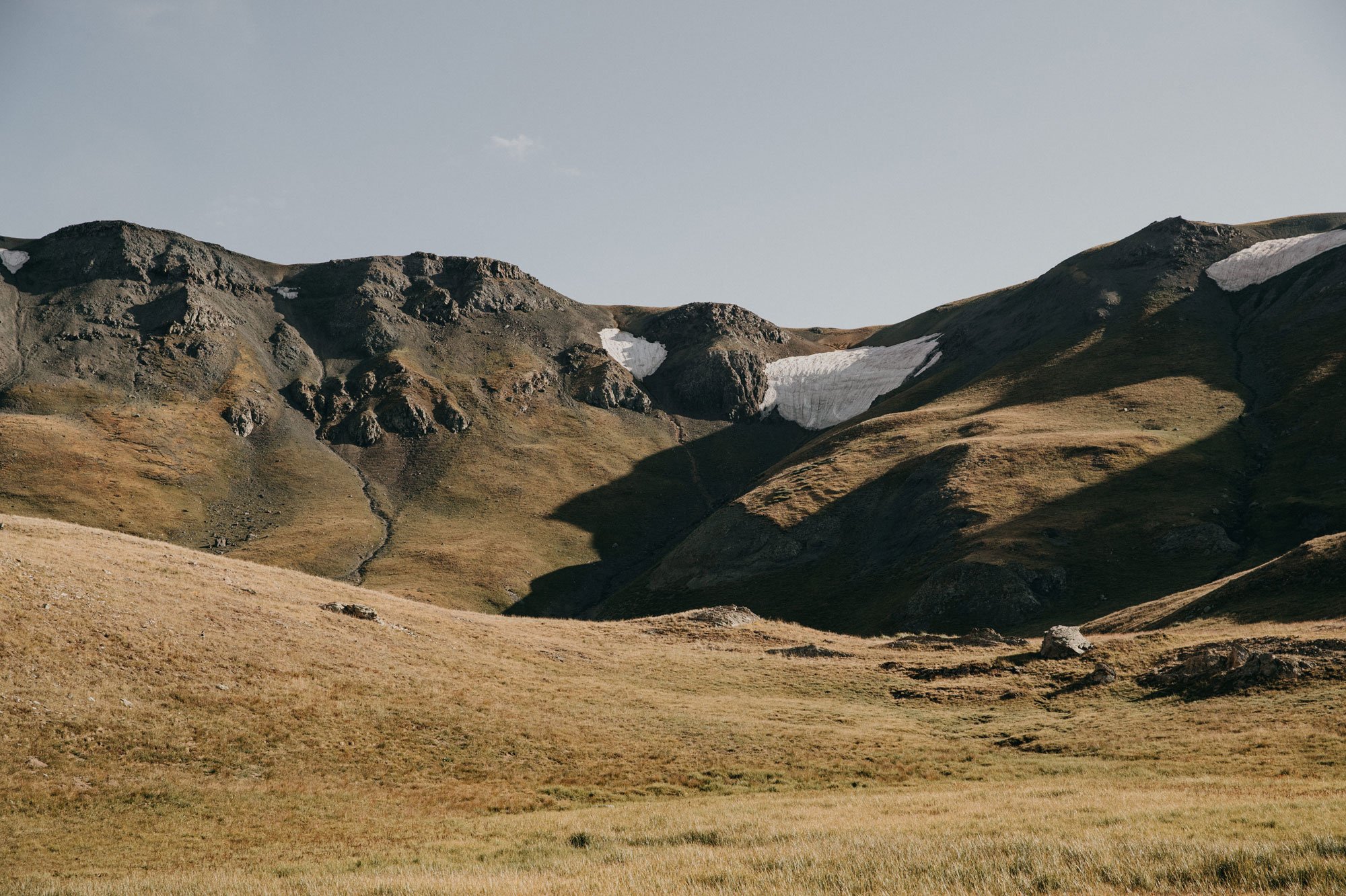 A landscape of rolling grassy hills with rocky mountains in the background and patches of snow, under a clear sky in Colorado