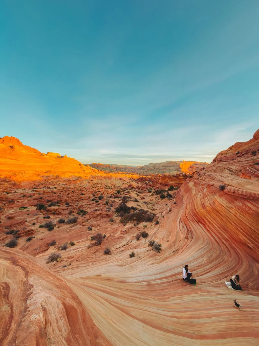 Two people sitting on colorful striped rock formations in a desert landscape under a clear blue sky.