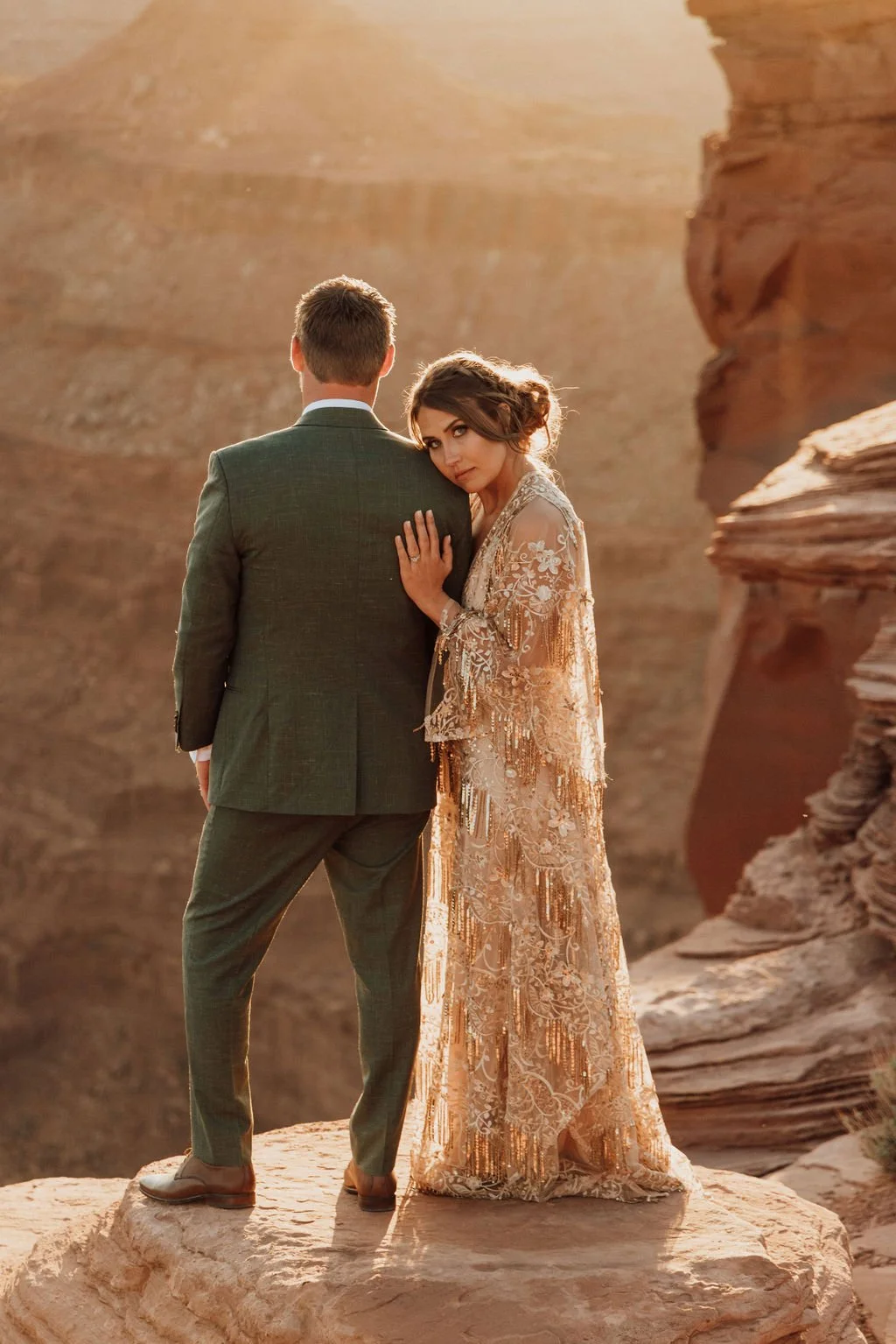 A couple stands on a rock in a desert canyon; the woman is wearing an ornate, gold-embellished dress and leans into the man's back, who is dressed in a suit, with her hand on his shoulder and her head resting on his shoulder.