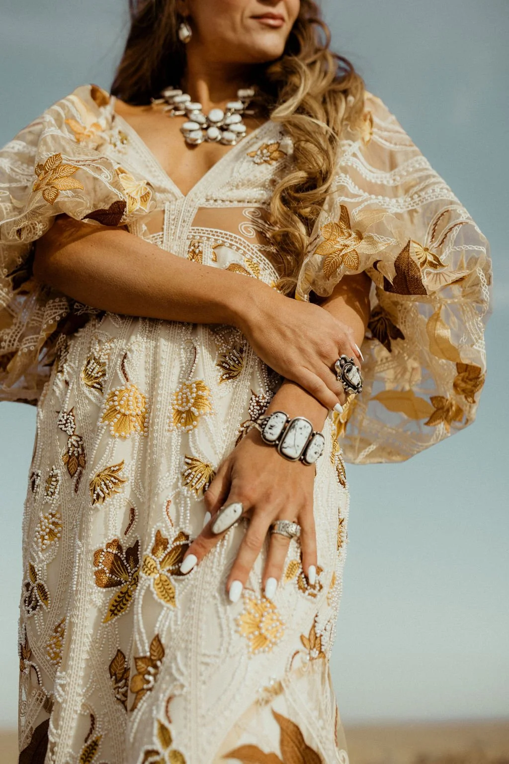 Close-up of a woman wearing an ornate, cream-colored dress with gold and brown embroidery, adorned with jewelry including rings, a bracelet with turquoise stones, and a pearl necklace, standing outdoors against a blue sky.
