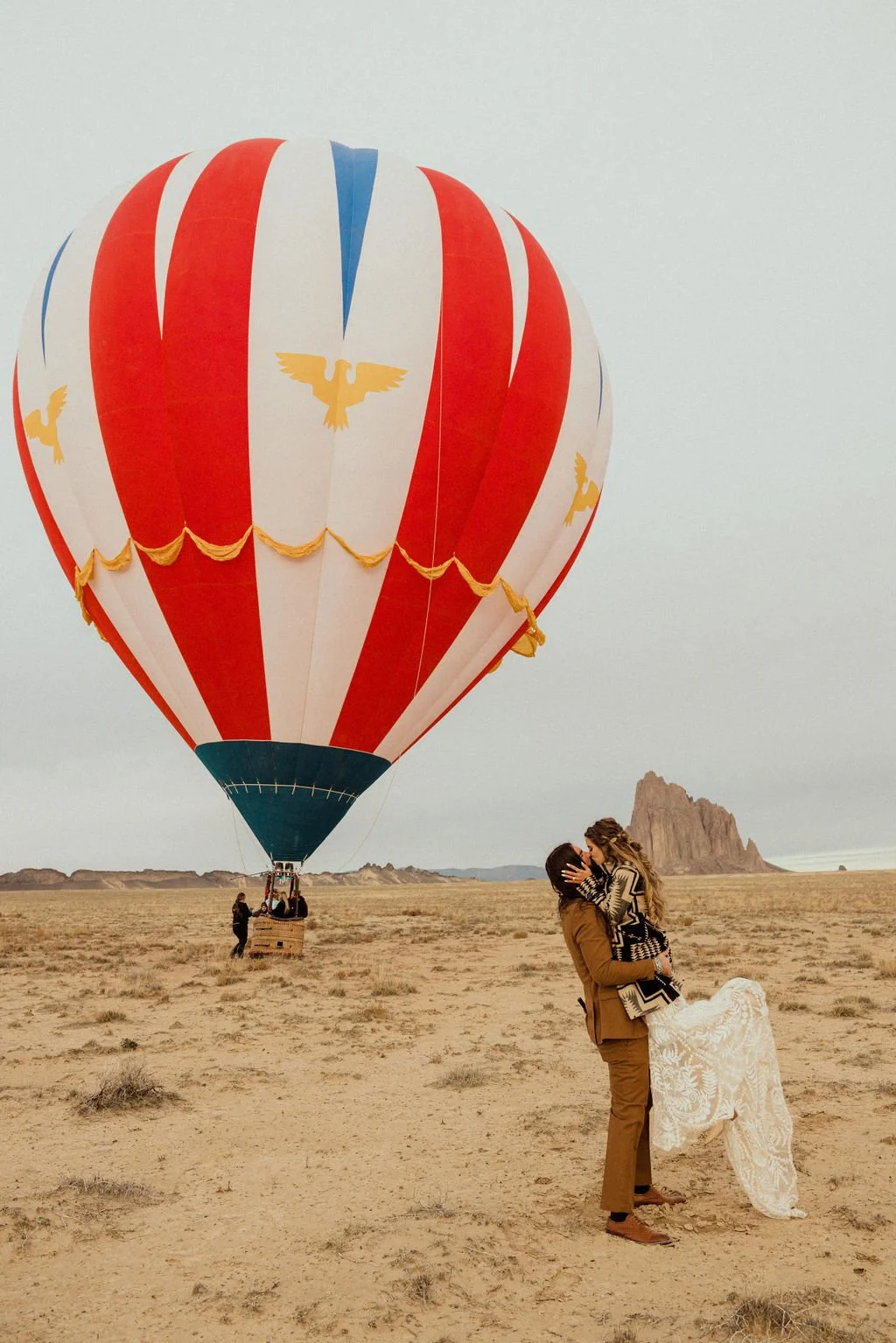 A couple sharing a kiss in a desert landscape with a large red and white hot air balloon featuring a yellow bird emblem in the background.