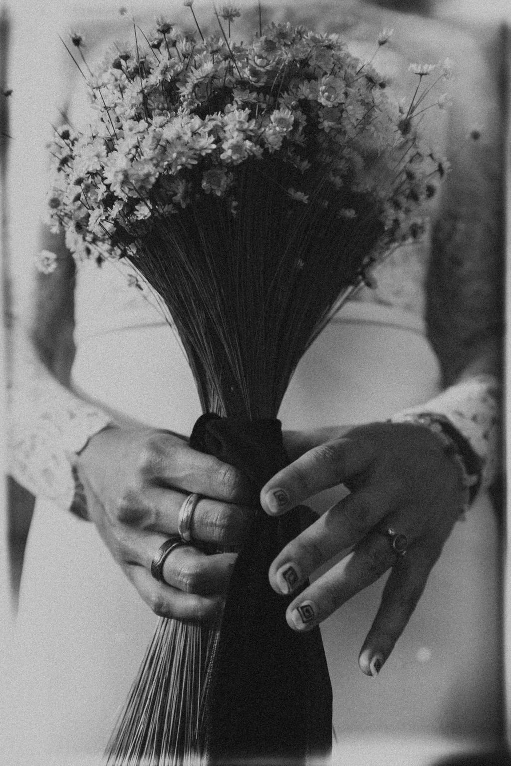 Close-up of a person holding a bouquet of small flowers wrapped with a black ribbon, showing hands with rings and decorated nails.