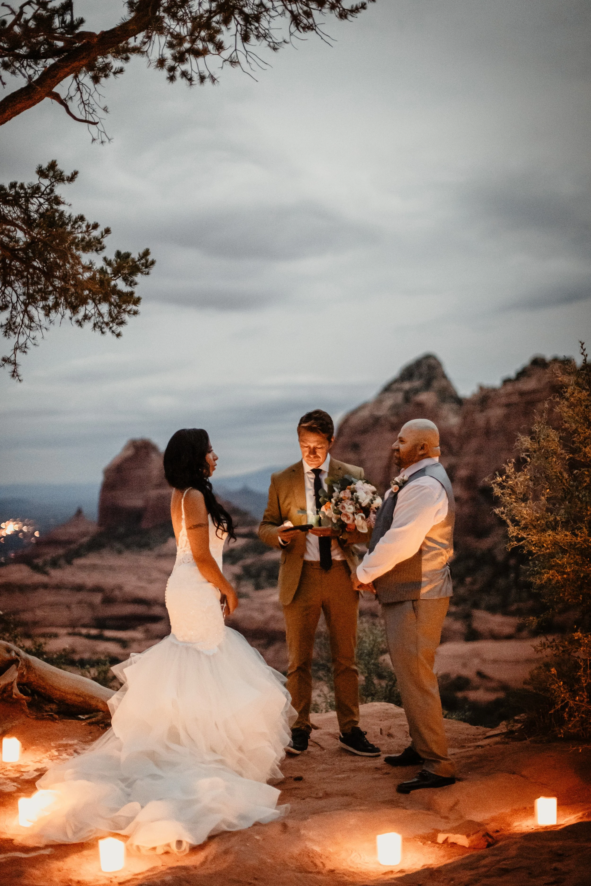 A wedding ceremony taking place outdoors at sunset, with a bride in a white gown, a groom in a vest, and a officiant holding a bouquet of flowers, surrounded by candles on the ground and rocky mesas in the background.
