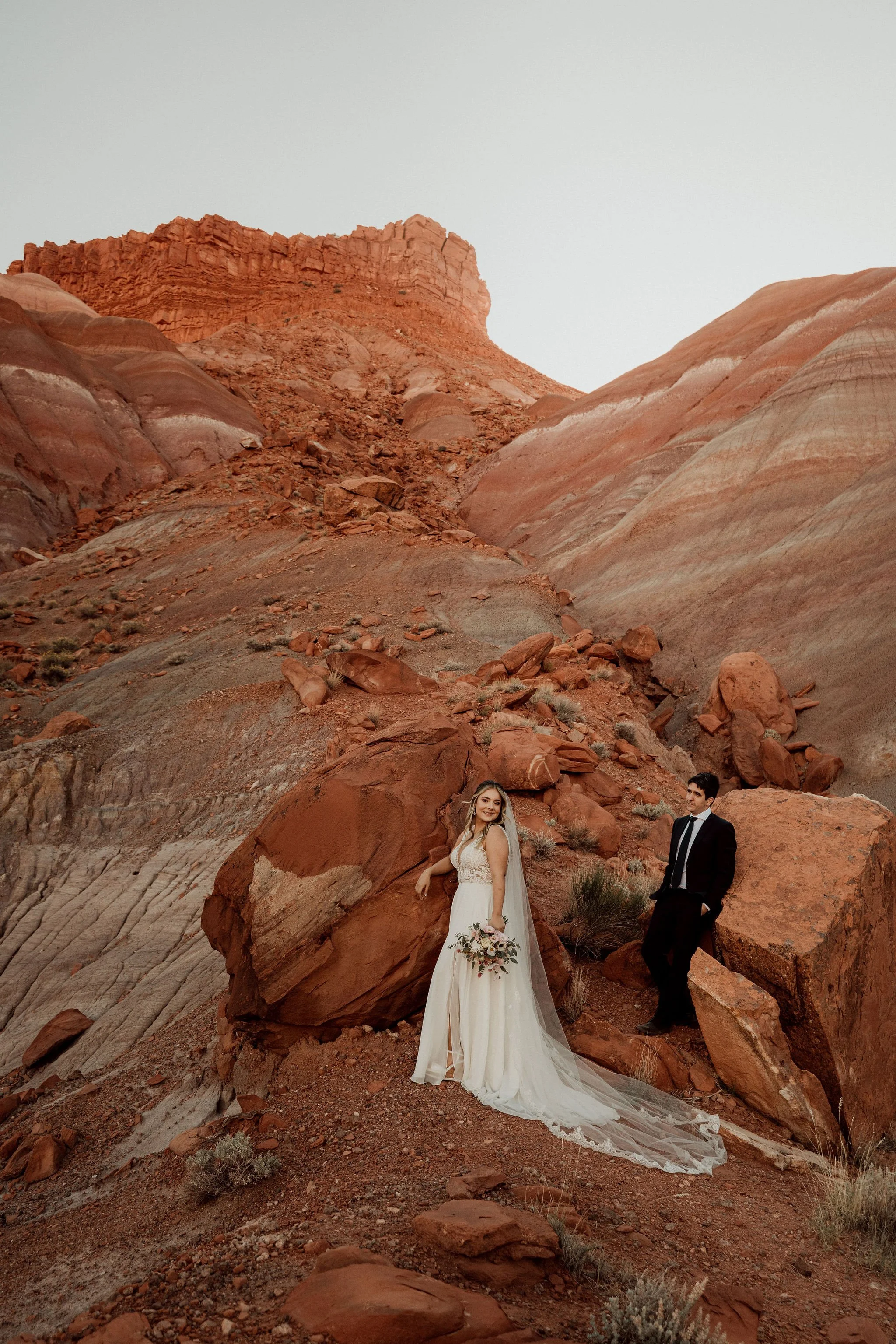 Bride in a white wedding gown holding a bouquet, and groom in a black suit with a tie, standing among red rocks and cliffs in a desert landscape.