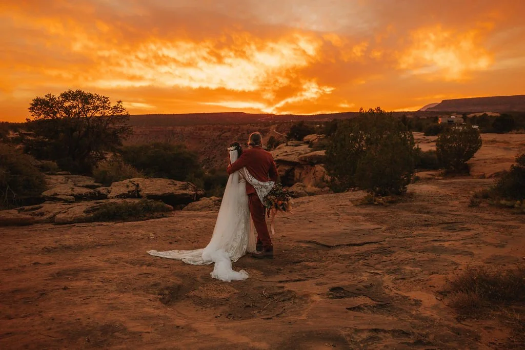 A couple embracing at sunset in the Arizona desert landscape, with rocky terrain and scattered bushes.