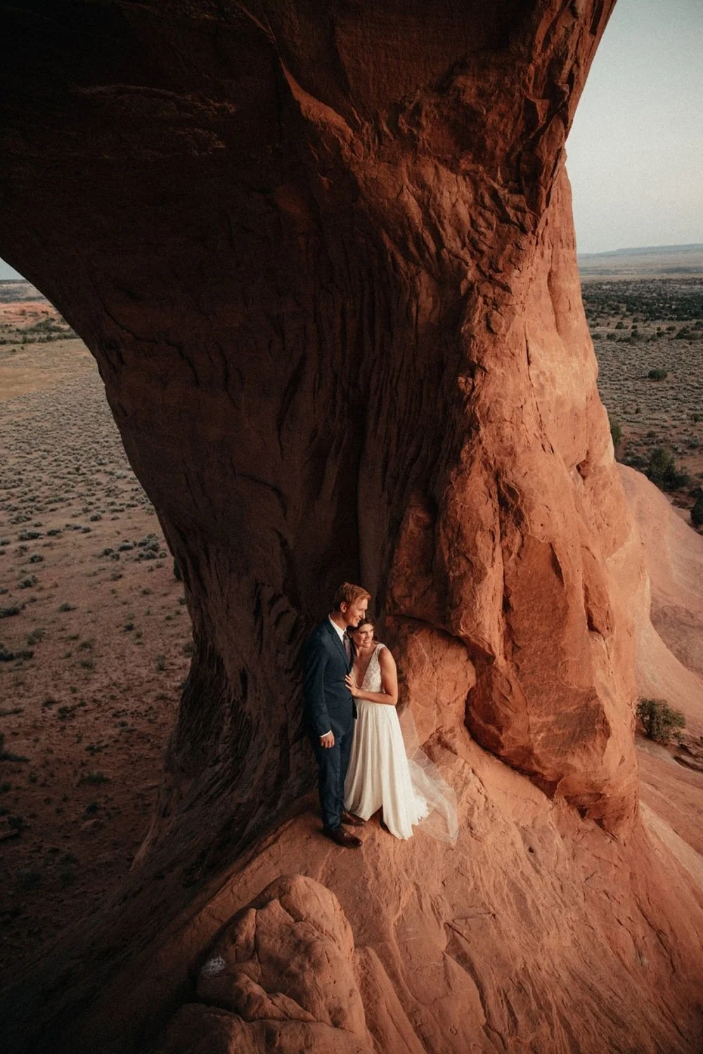 A bride and groom stand together at sunset on a red rock formation in the Utah desert.