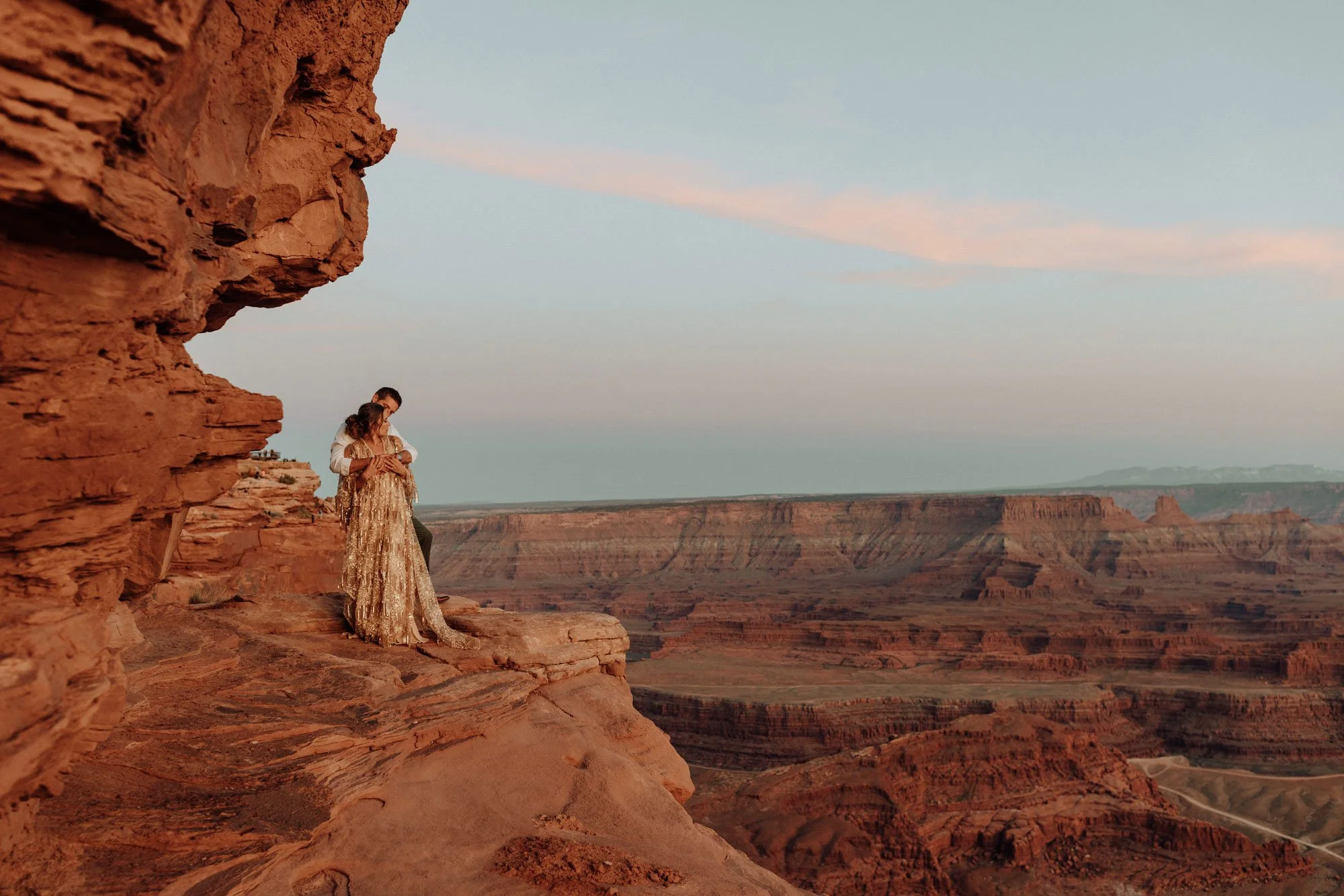 A couple stands on a rocky ledge overlooking the desert at sunset, embracing each other with the expansive canyon landscape behind them.