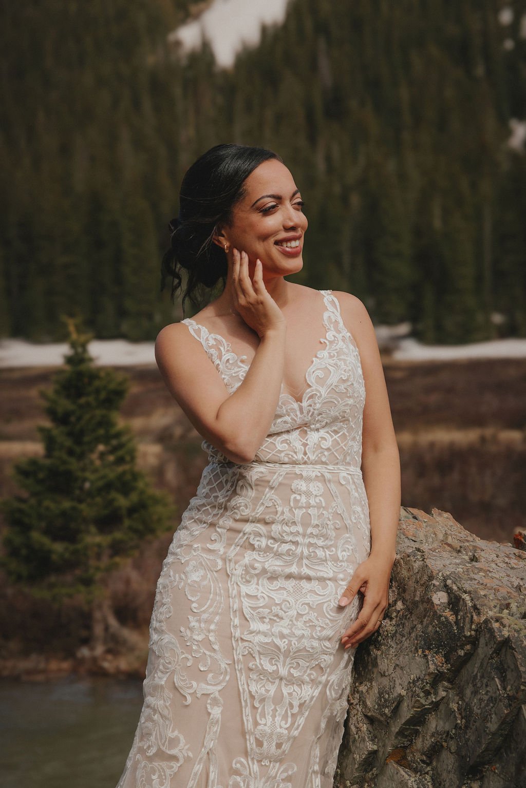 A woman in a white lace dress standing outdoors next to a rock, smiling with her hand touching her face, with a forest and river in the background.