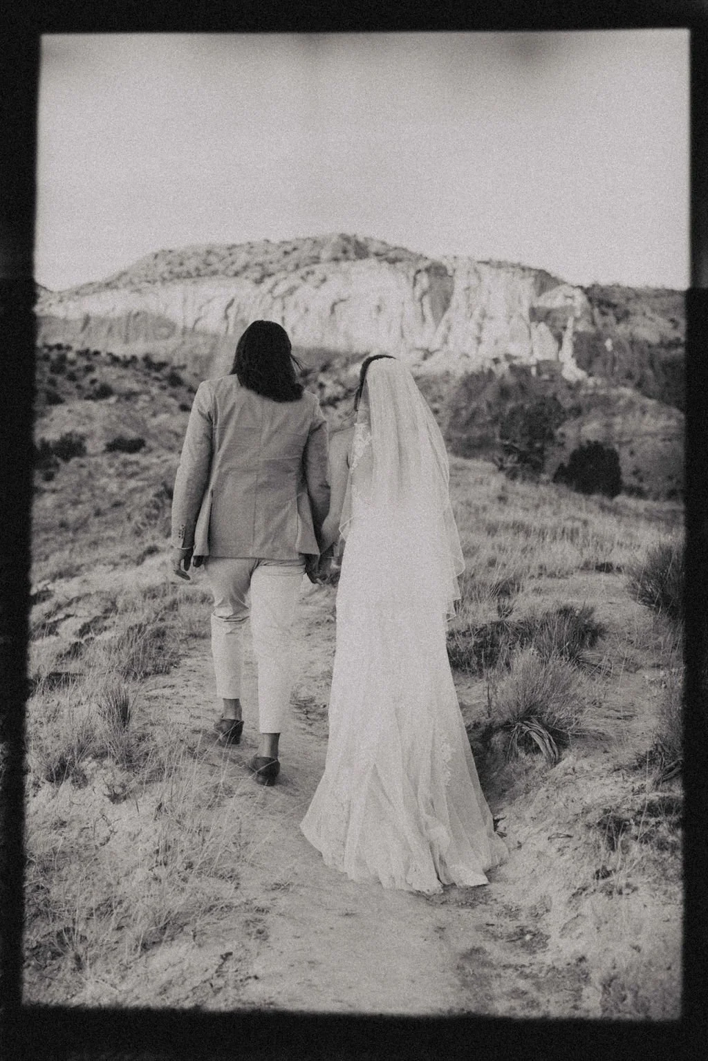 Two women, one in a wedding dress and the other in a suit, hold hands and walk along a dirt path through a desert landscape with rocky hills in the background, black and white photograph.