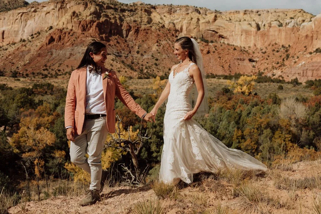 A woman in a wedding dress and a person in a peach blazer holding hands in a desert landscape with red rock formations and colorful foliage.
