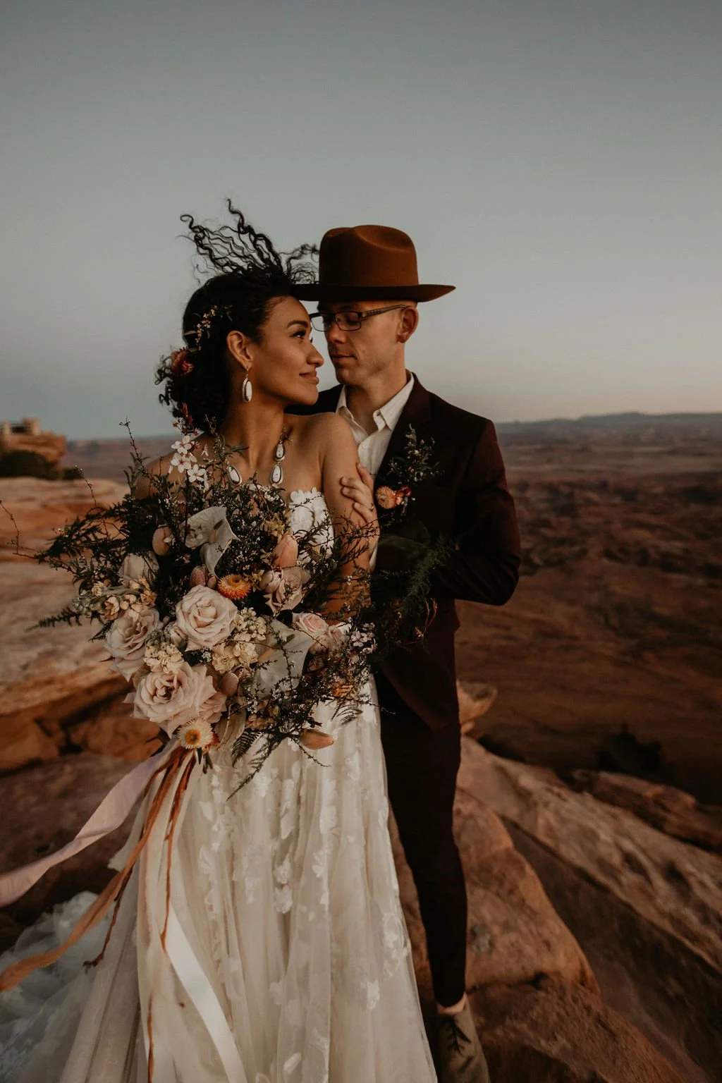 A couple dressed in wedding attire standing on rocky terrain during sunset, with a scenic landscape in the background. The woman is holding a large bouquet of flowers, and both appear to be gazing lovingly at each other.