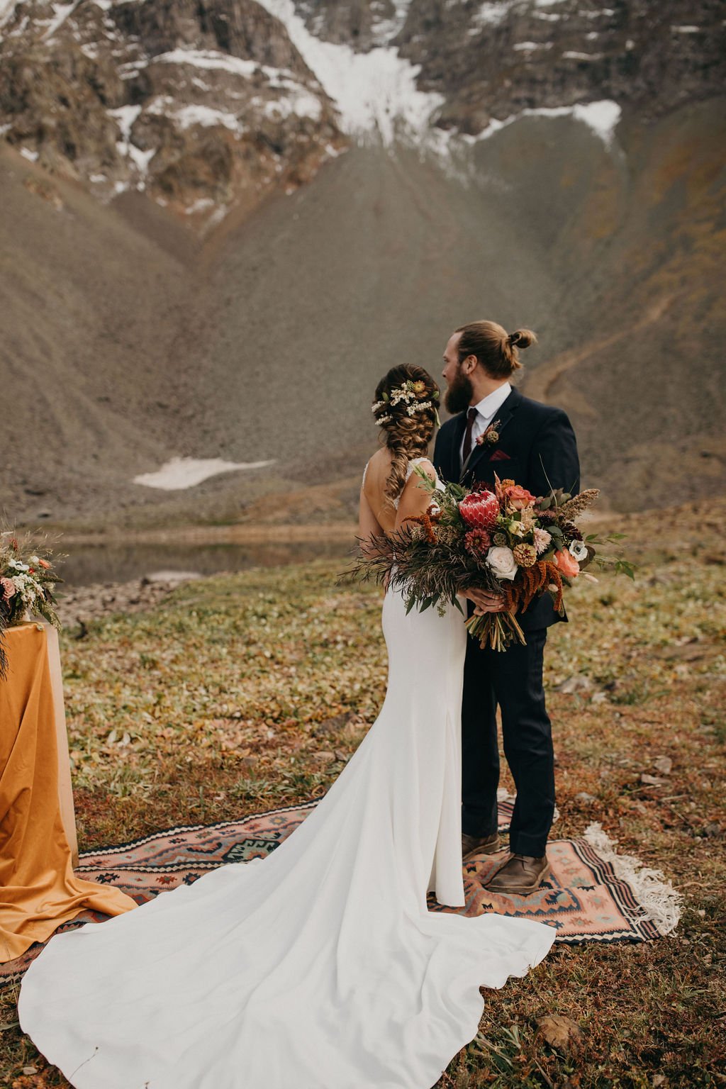 A wedding ceremony featuring a bride and groom standing on a patterned rug outdoors in a mountainous area with snow patches. The bride wears a white dress with a long train and floral hair accessories, holding a large bouquet of flowers. The groom is