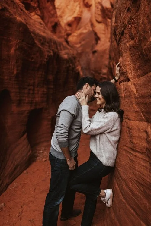 A couple sharing an intimate moment in a narrow red rock canyon, with the man leaning against the rock and the woman leaning in close, smiling.