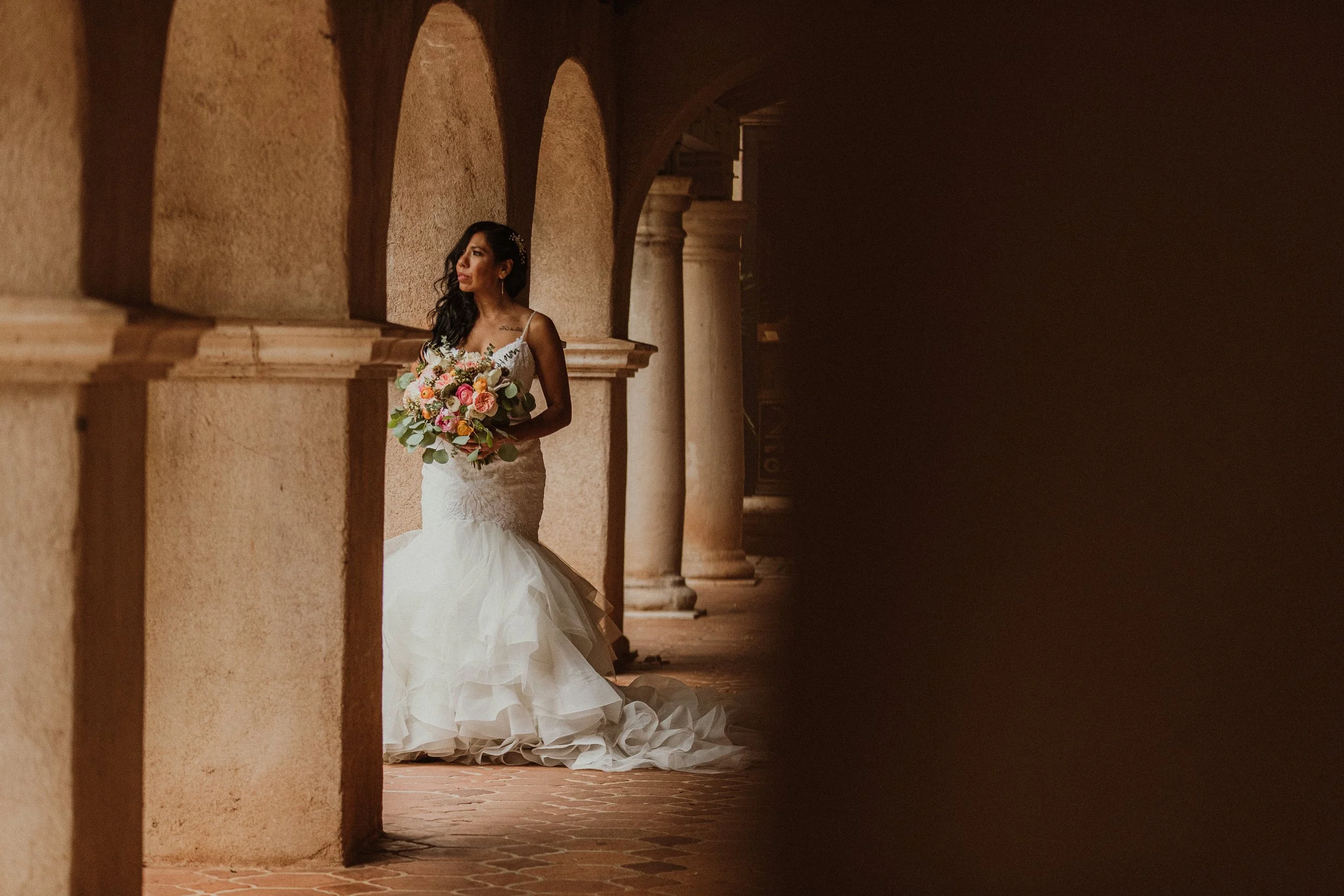 A bride in a white wedding gown holding a bouquet of pink, peach, and white flowers, standing in an arched corridor with stone columns.
