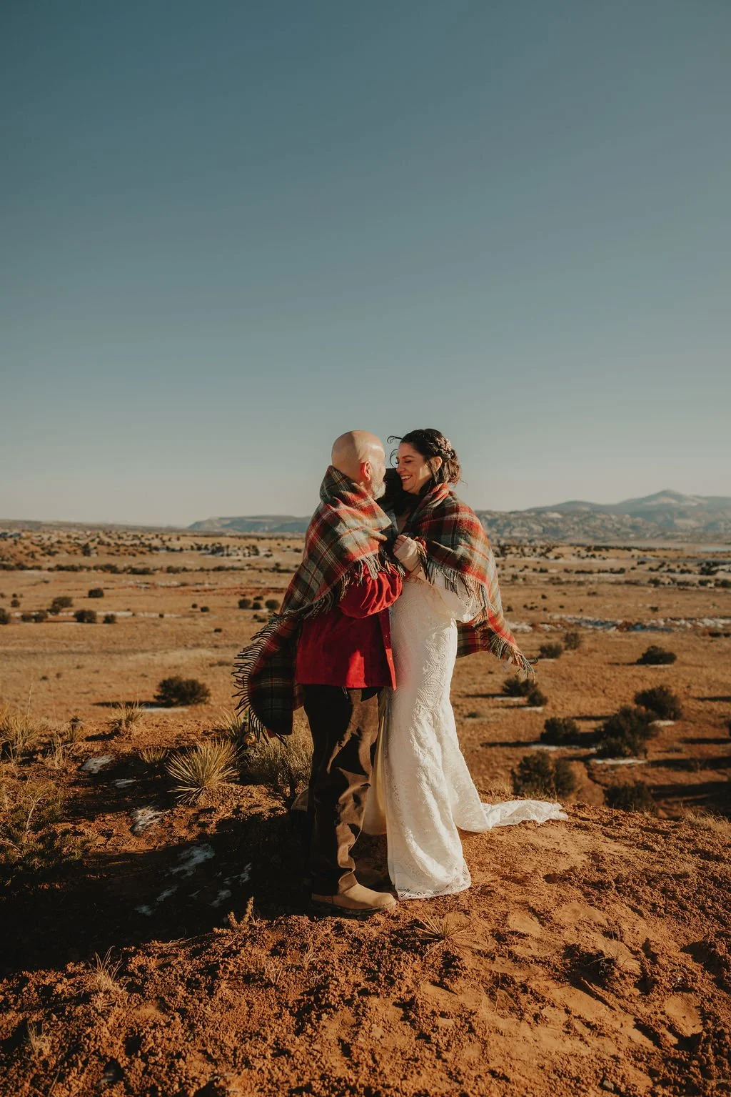 A couple dressed as a bride and groom stands in a desert landscape wrapped in plaid blankets, smiling at each other with mountains in the background.