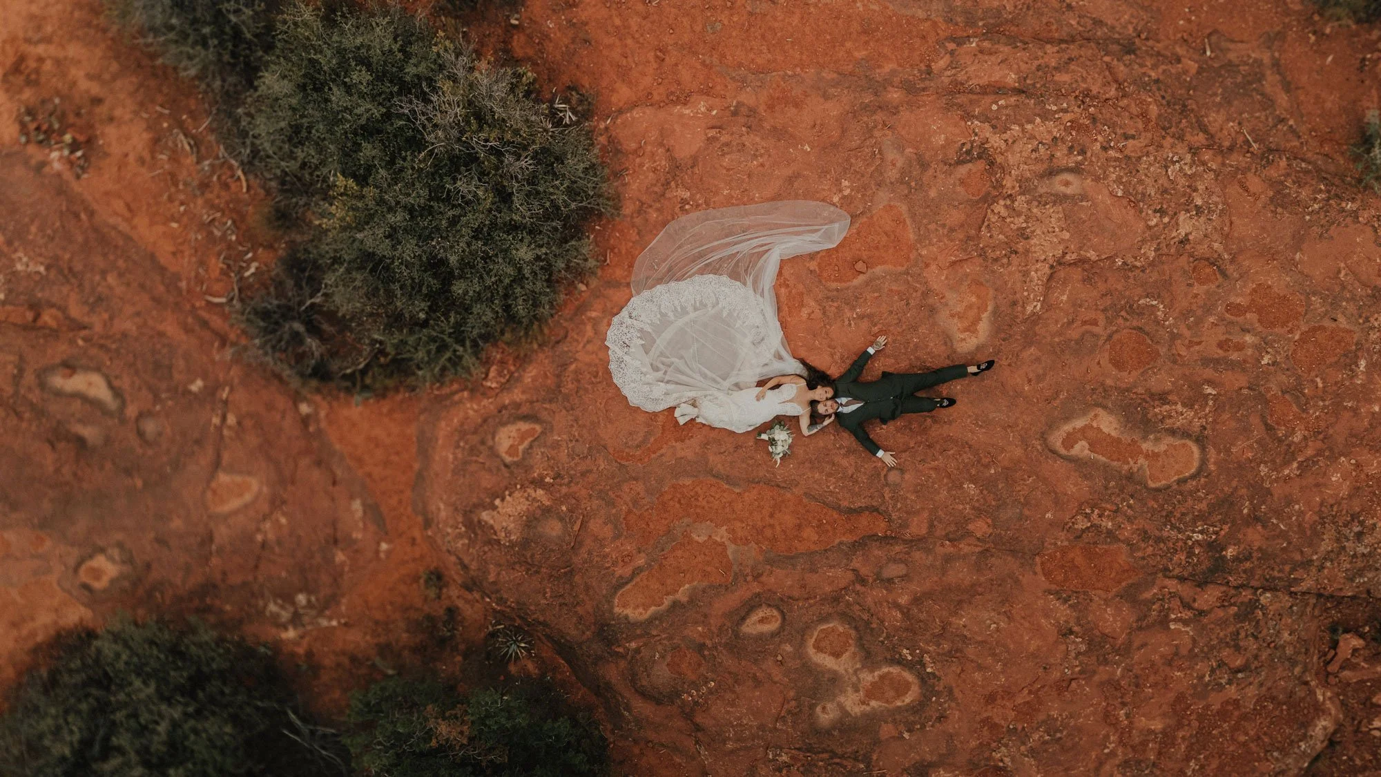 A bride and groom lying on red rocky ground in a desert-like landscape, with the bride in a white wedding dress and veil, and the groom in a dark suit, both gesturing with their arms outstretched.
