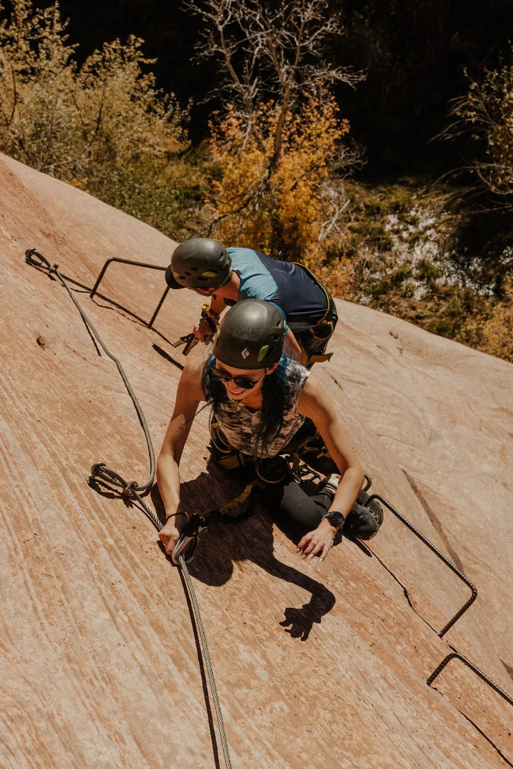 Two people rock climbing on a steep, sandy-colored rock face with safety harnesses and helmets, with trees in autumn colors in the background.