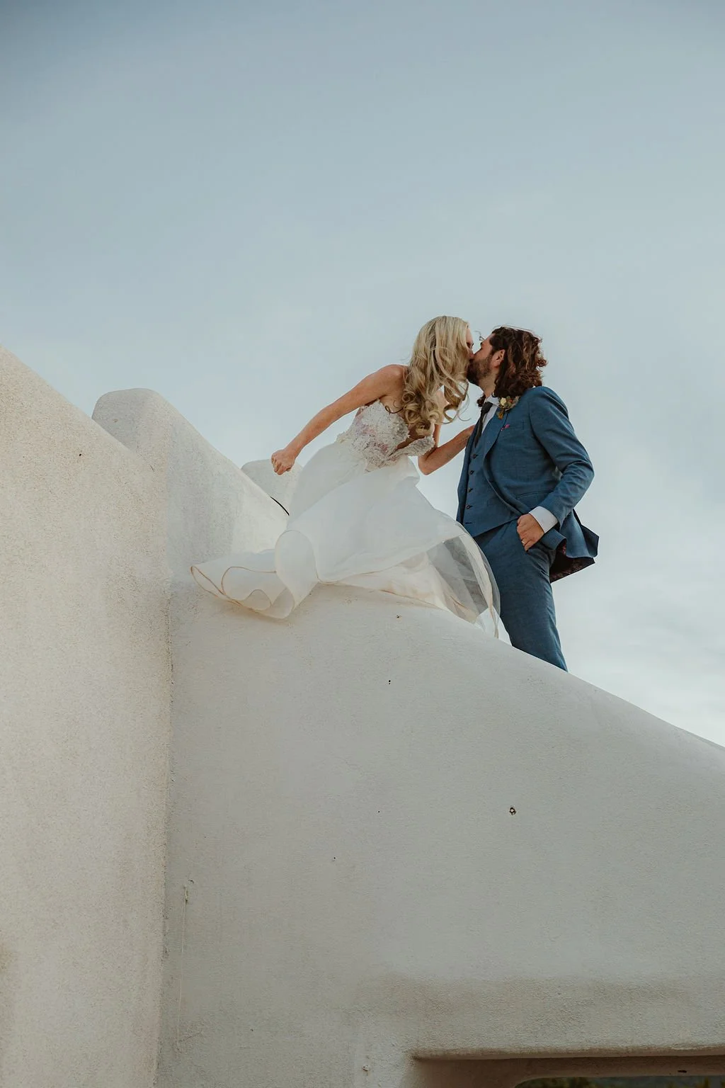 A bride and groom kiss on a rooftop against a pale sky, with the bride sitting on the edge and the groom standing nearby.