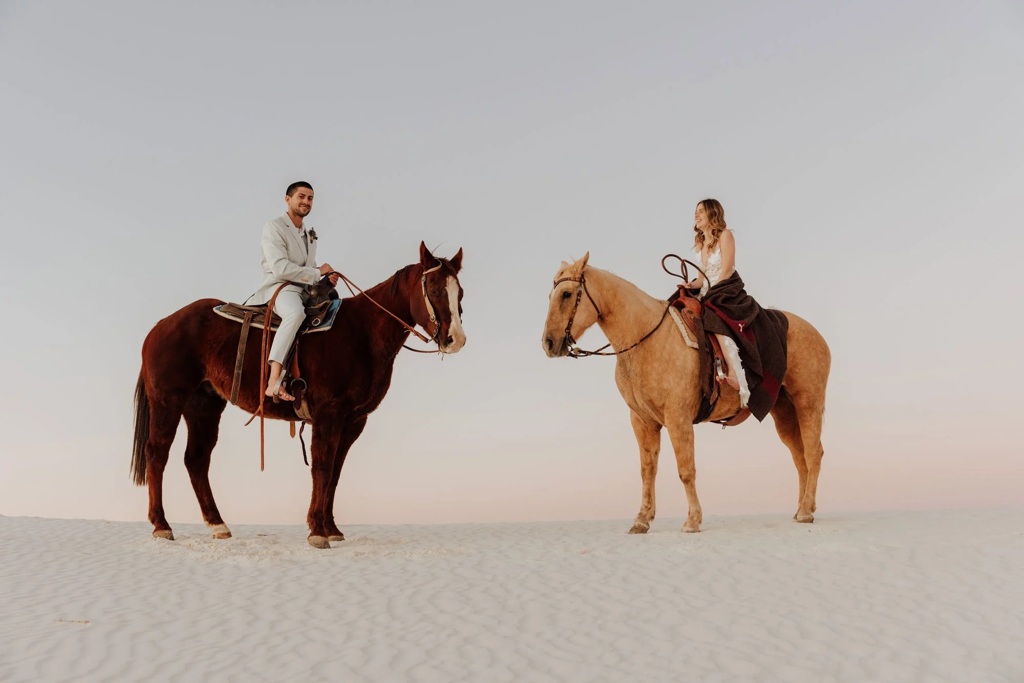 A man and a woman riding horses on a sandy surface under an open sky, facing each other and smiling.