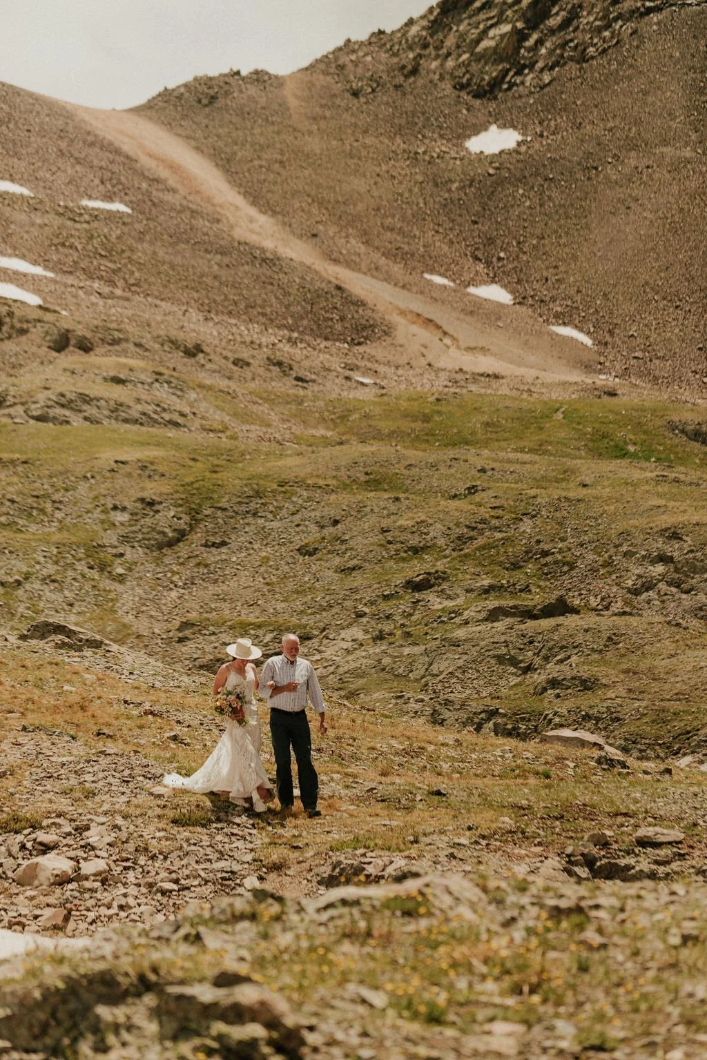 A bride and her father walking together on a rocky trail in a mountainous area with patches of snow and green grass in Colorado