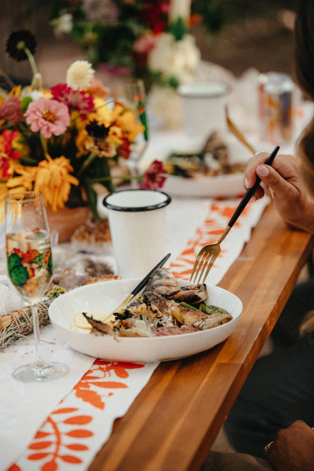 Person using a fork to eat fish from a white bowl on a decorated dining table with flowers, wine glass, paper cup, and a festive table runner.