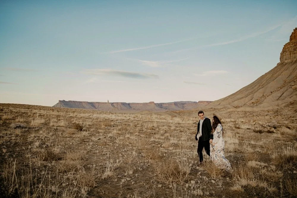 A man and woman walking together in a barren desert landscape with plateaus and hills in the background under a partly cloudy sky.