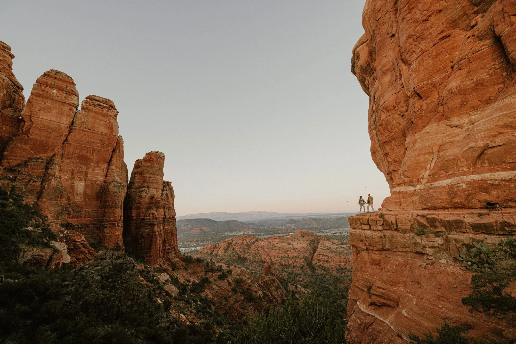 Two people holding hands on a small ledge of red rock cliffs in a desert canyon with a distant view of layered rock formations and a clear sky.