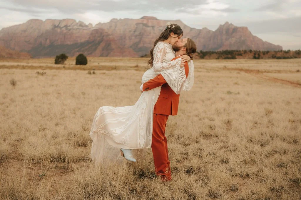 A couple in wedding attire embracing in an open field with mountains in the background.