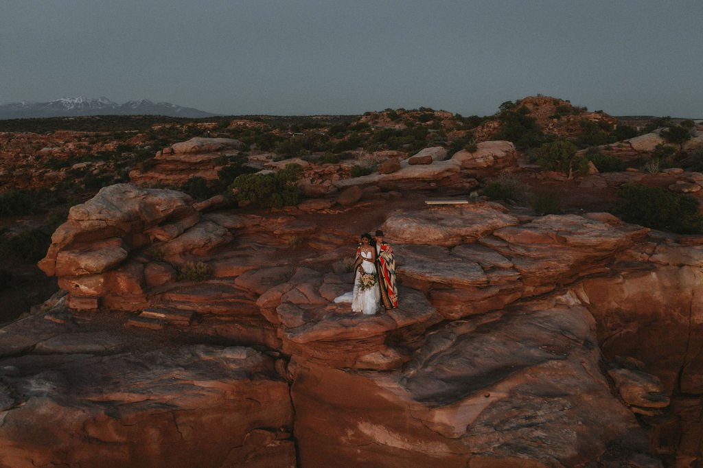 A bride and groom standing on large rocky terrain in a desert landscape with mountains in the background.