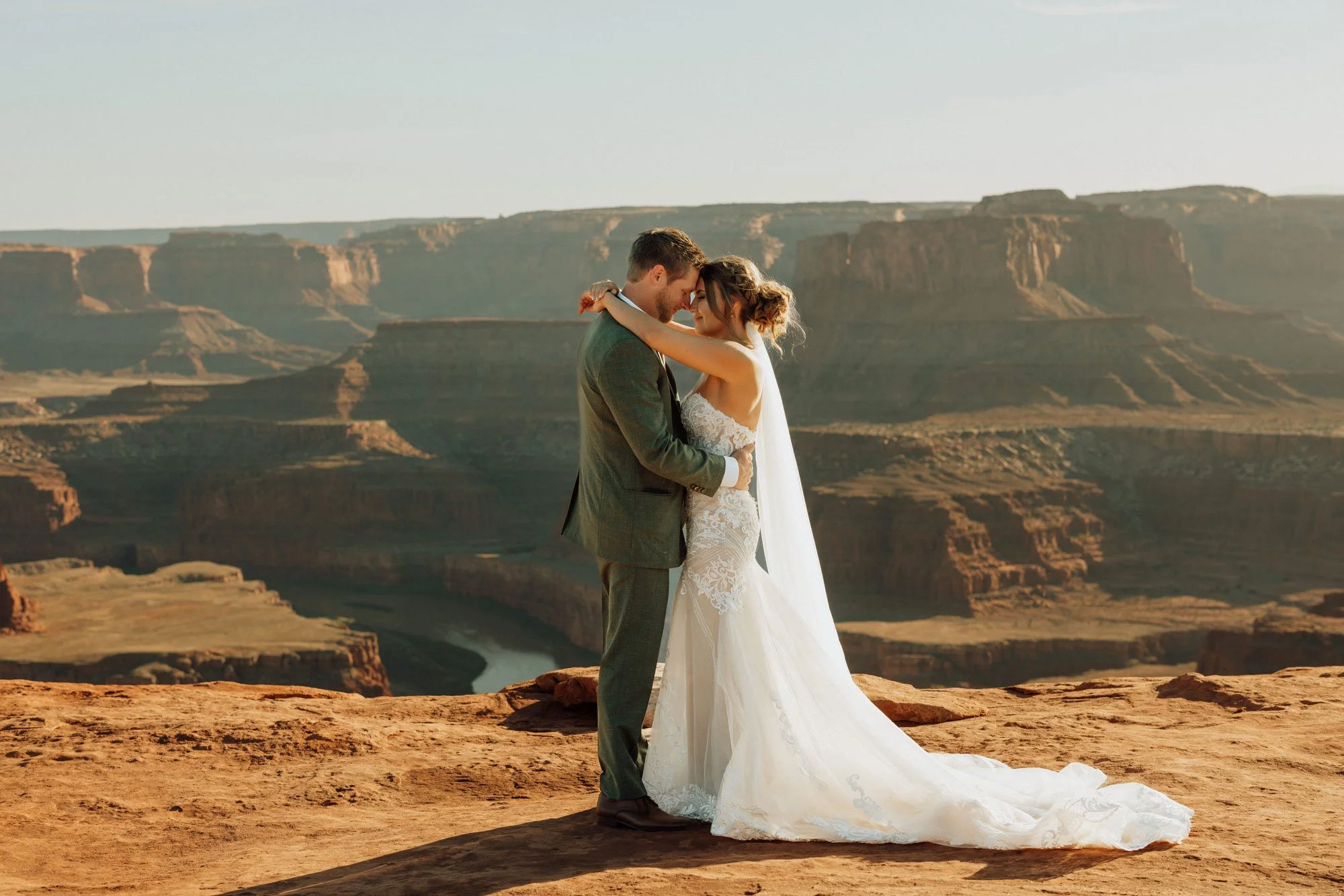 A bride and groom embrace on the edge of a canyon, with a river winding through the landscape in the background.